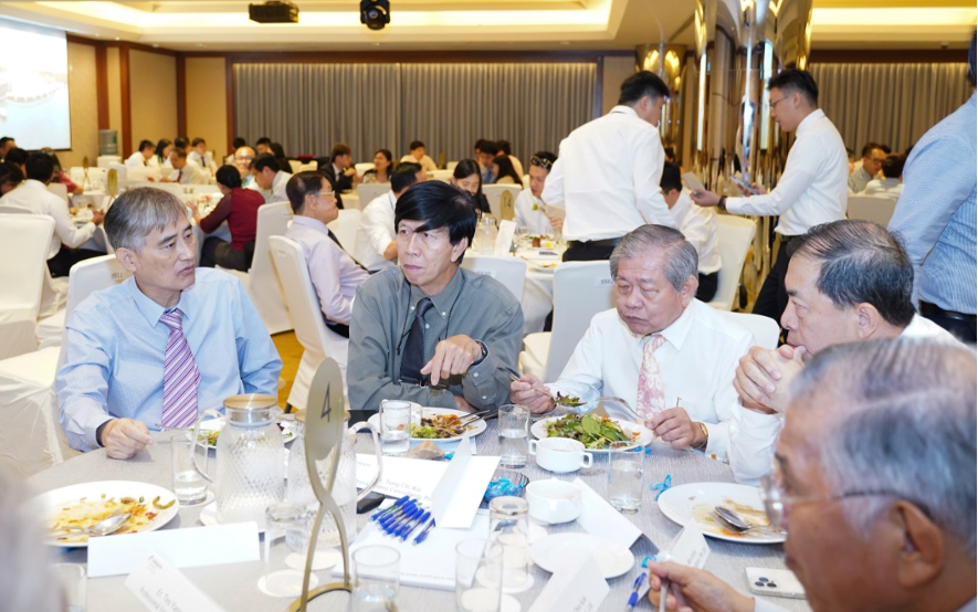 Several older men in formal attire sit around a round dining table eating and conversing in a busy banquet hall.