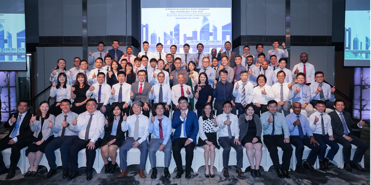 Large group of formally dressed professionals posing for a photo, many giving thumbs up, with a presentation screen behind them.