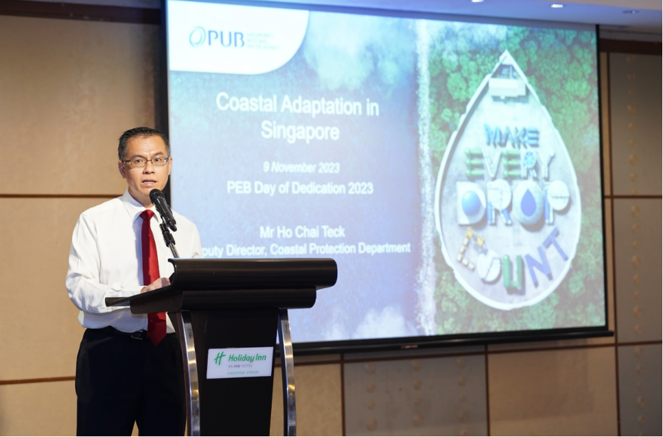 Man in white shirt and red tie speaks at a Holiday Inn podium before a PUB "Coastal Adaptation in Singapore" slide.