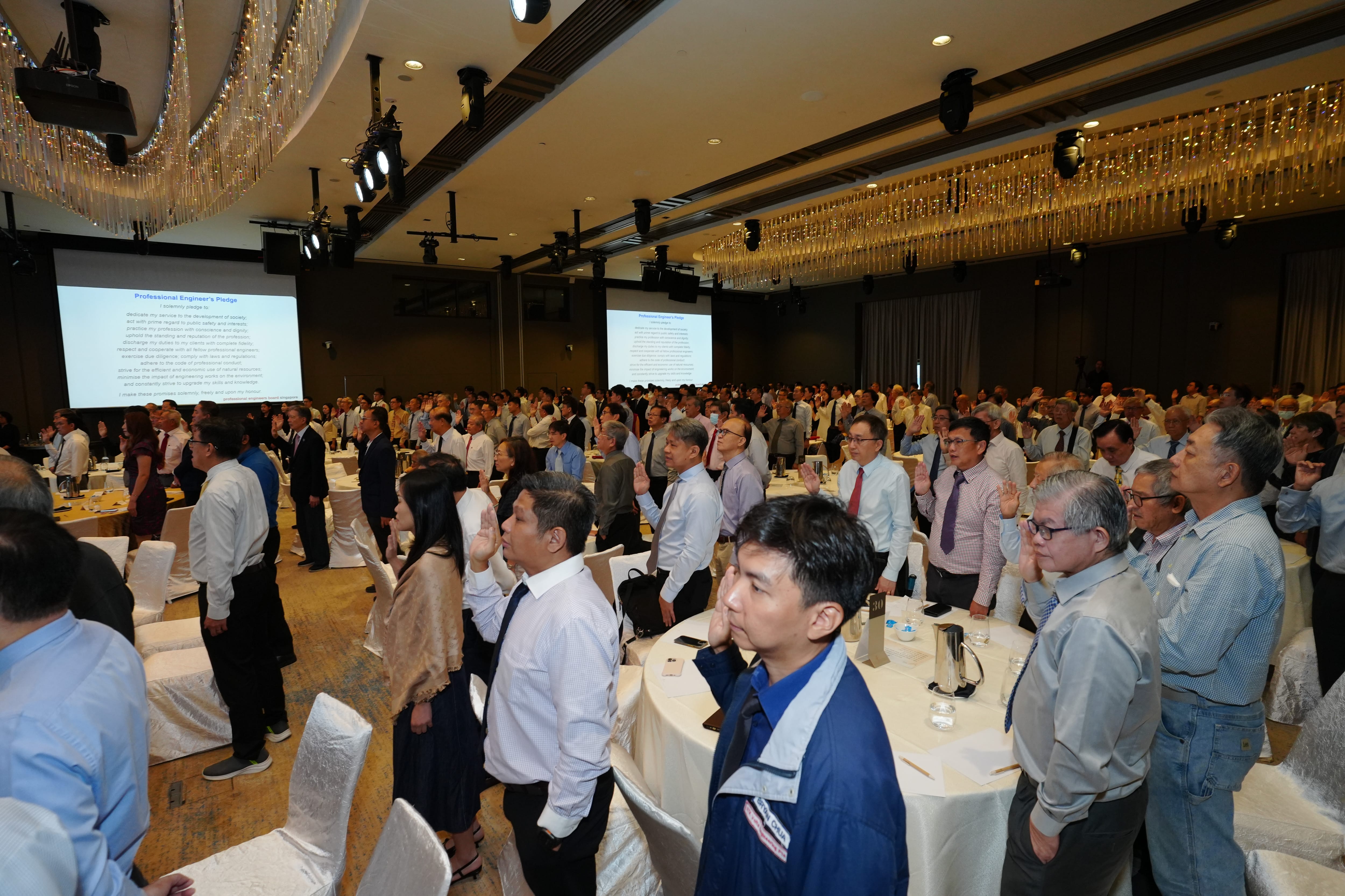 A large group of people standing with right hands raised reciting a Professional Engineer's Pledge shown on screens in a ballroom.