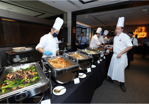 Two chefs in white uniforms and tall hats tend to a buffet line with multiple chafing dishes of food.