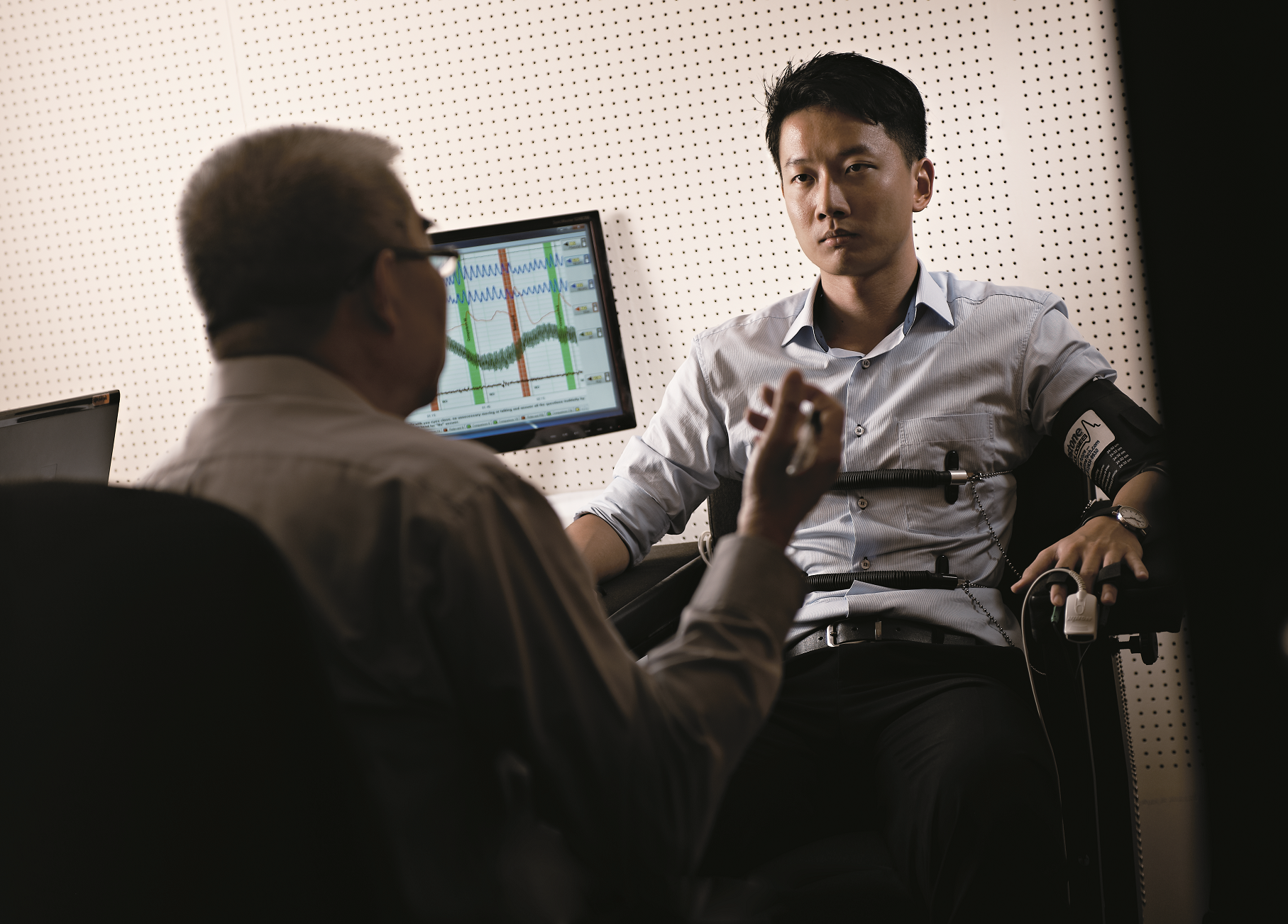 A young man connected to polygraph sensors sits facing an examiner, with a monitor showing physiological readings behind them.