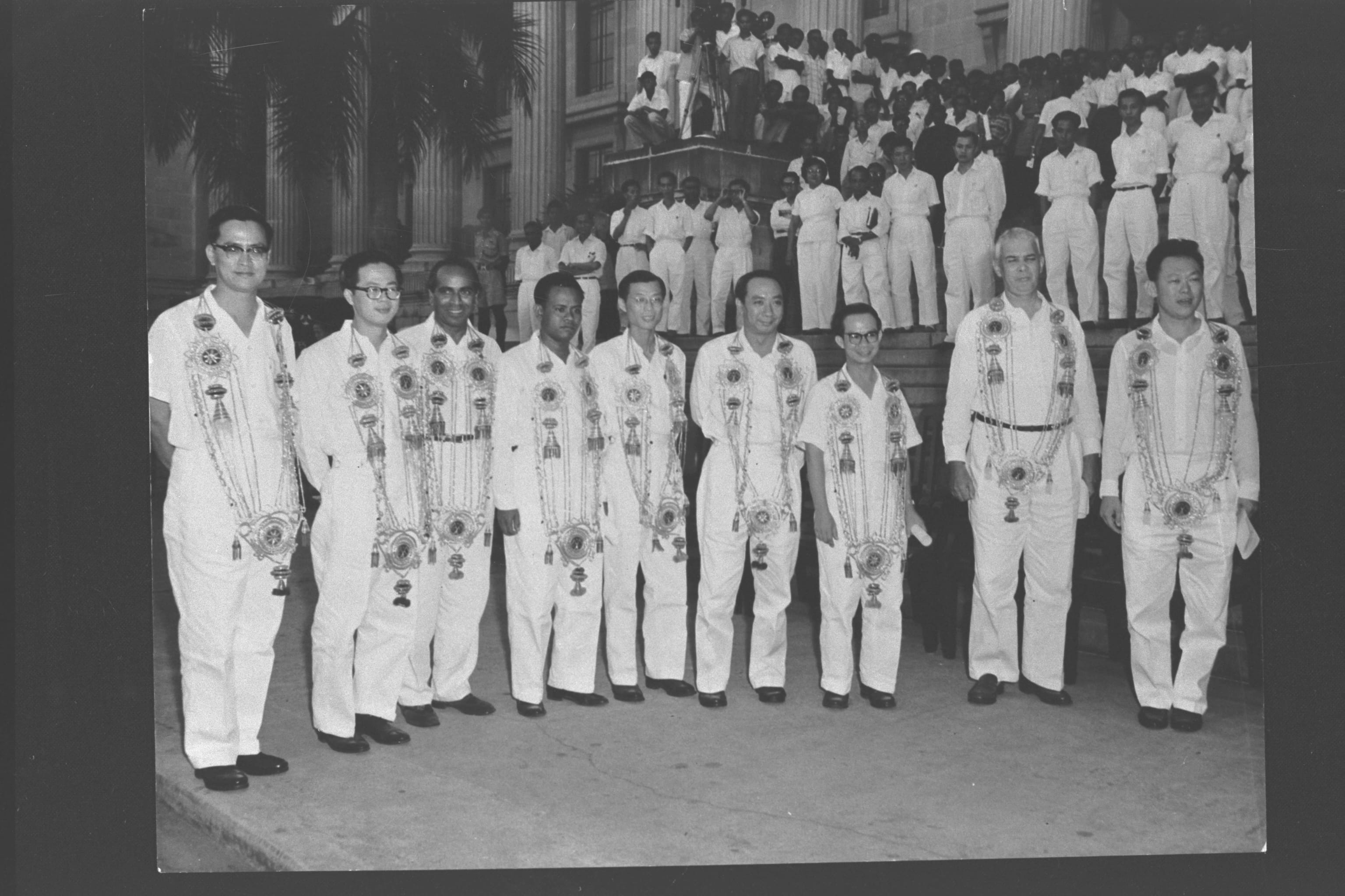 Eight men in white uniforms wearing ornate ceremonial necklaces stand in a row before a large crowd on steps.