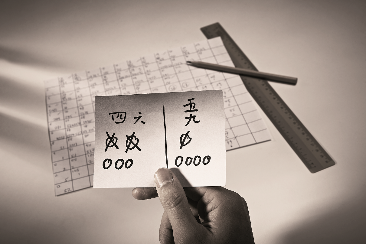 A hand holds a card with Chinese characters and tally marks, with a ruler, pencil, and grid paper in the background.