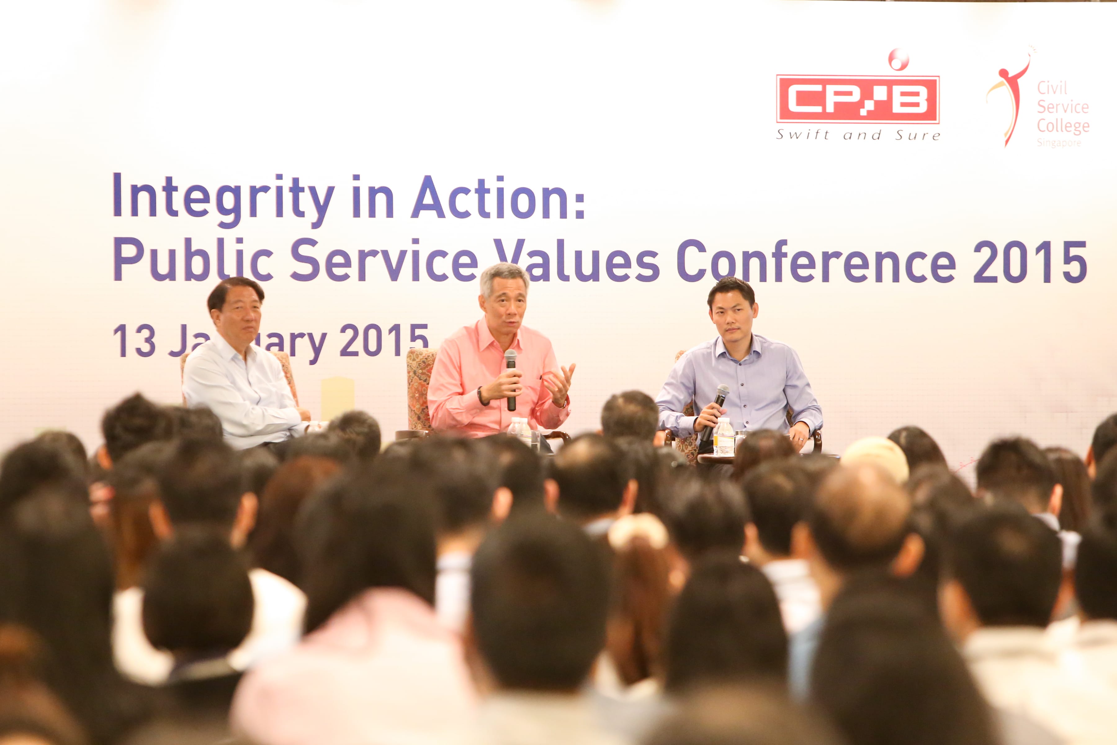 Three men sit on stage at the Public Service Values Conference 2015 on 13 January, speaking to a large audience.