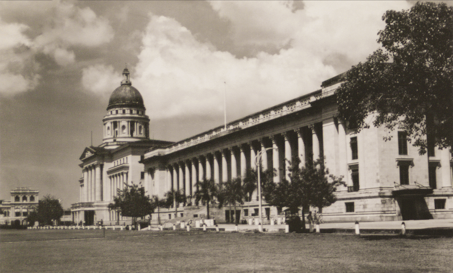 Vintage sepia photo of a grand colonial building with a dome, tall columns, open lawn, and trees under cloudy sky.