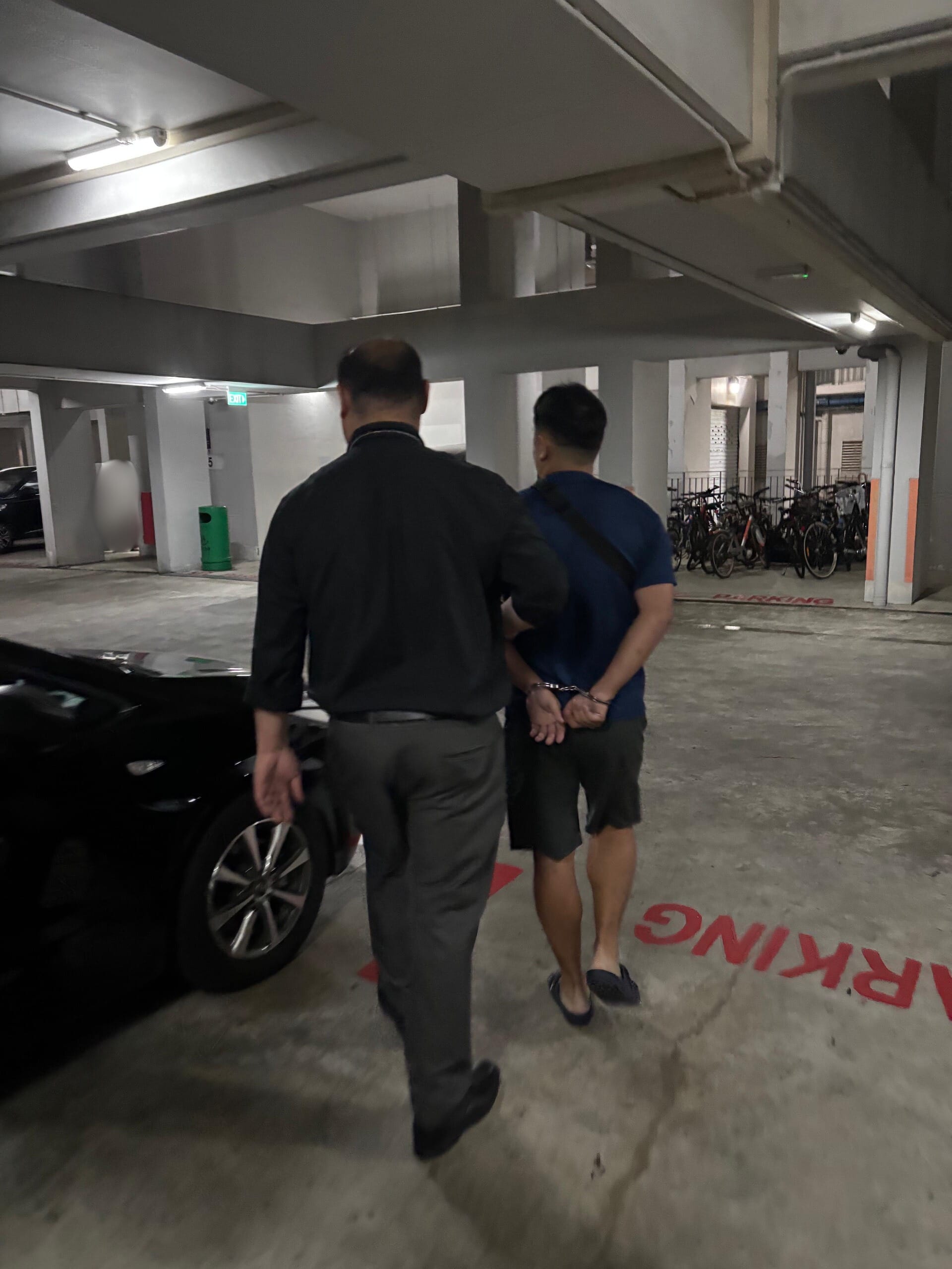 Two men walking in a HDB carpark, one in handcuffs being escorted by a CPIB officer in dark clothing.