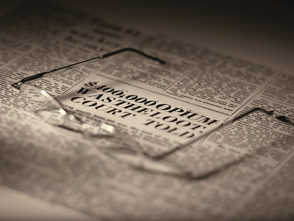 Wire-rimmed glasses resting on a sepia-toned newspaper with headline "$400,000 Opium Was The Loot Court Told."