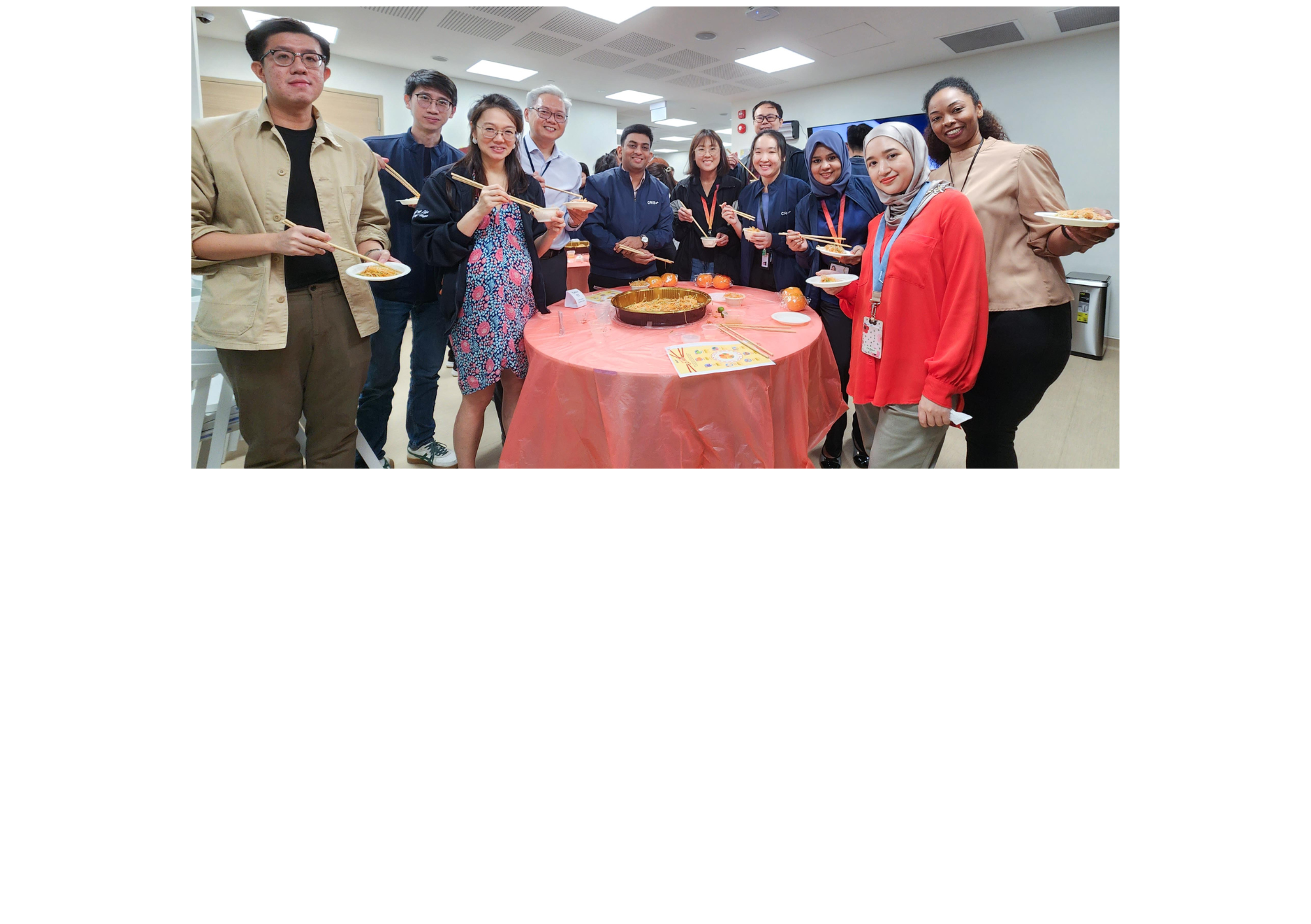 A group of people smiling around a red-covered table, holding chopsticks and plates of food indoors.