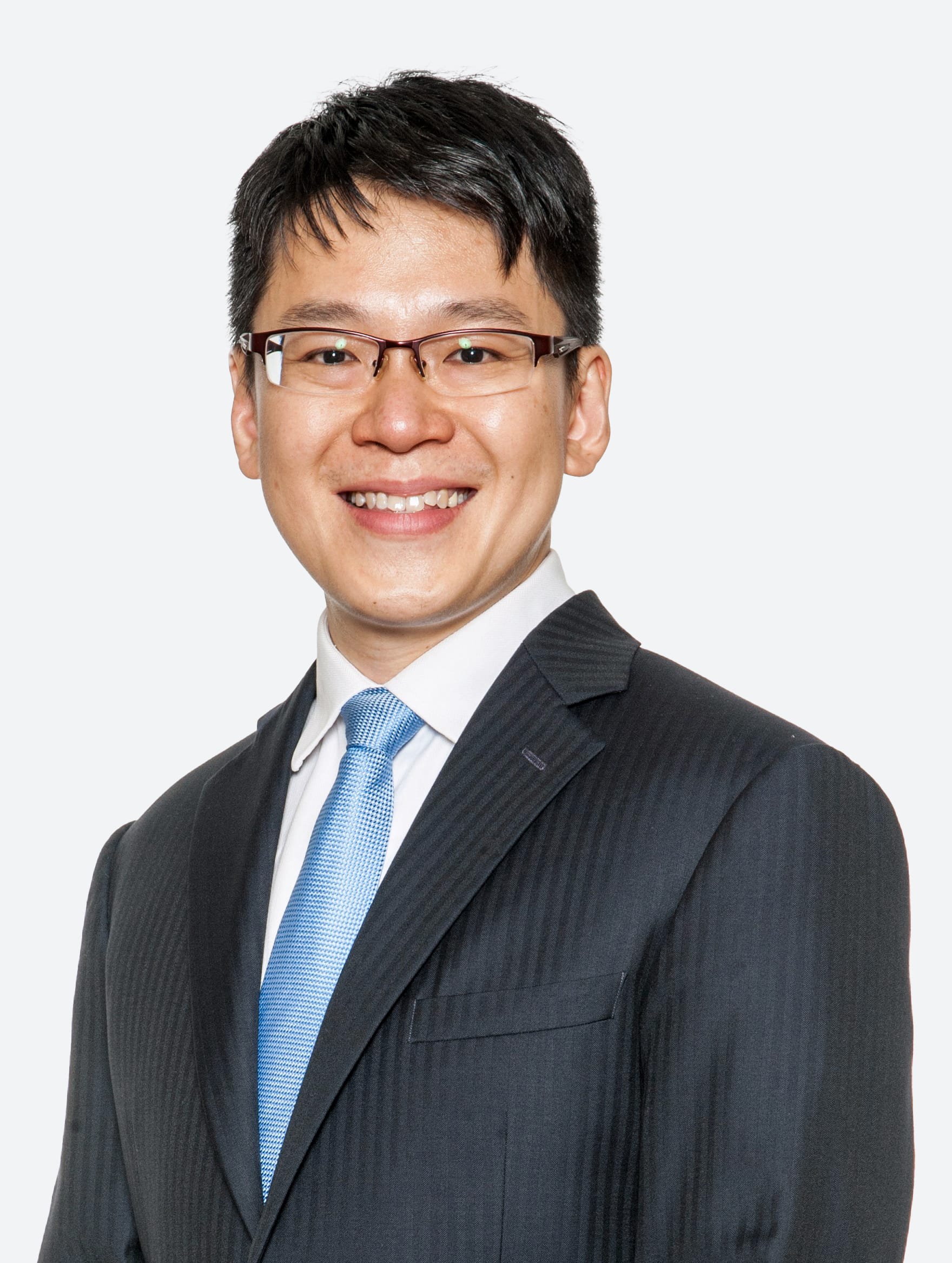 A smiling man wearing glasses, a dark suit, white shirt, and light blue tie against a pale grey background.