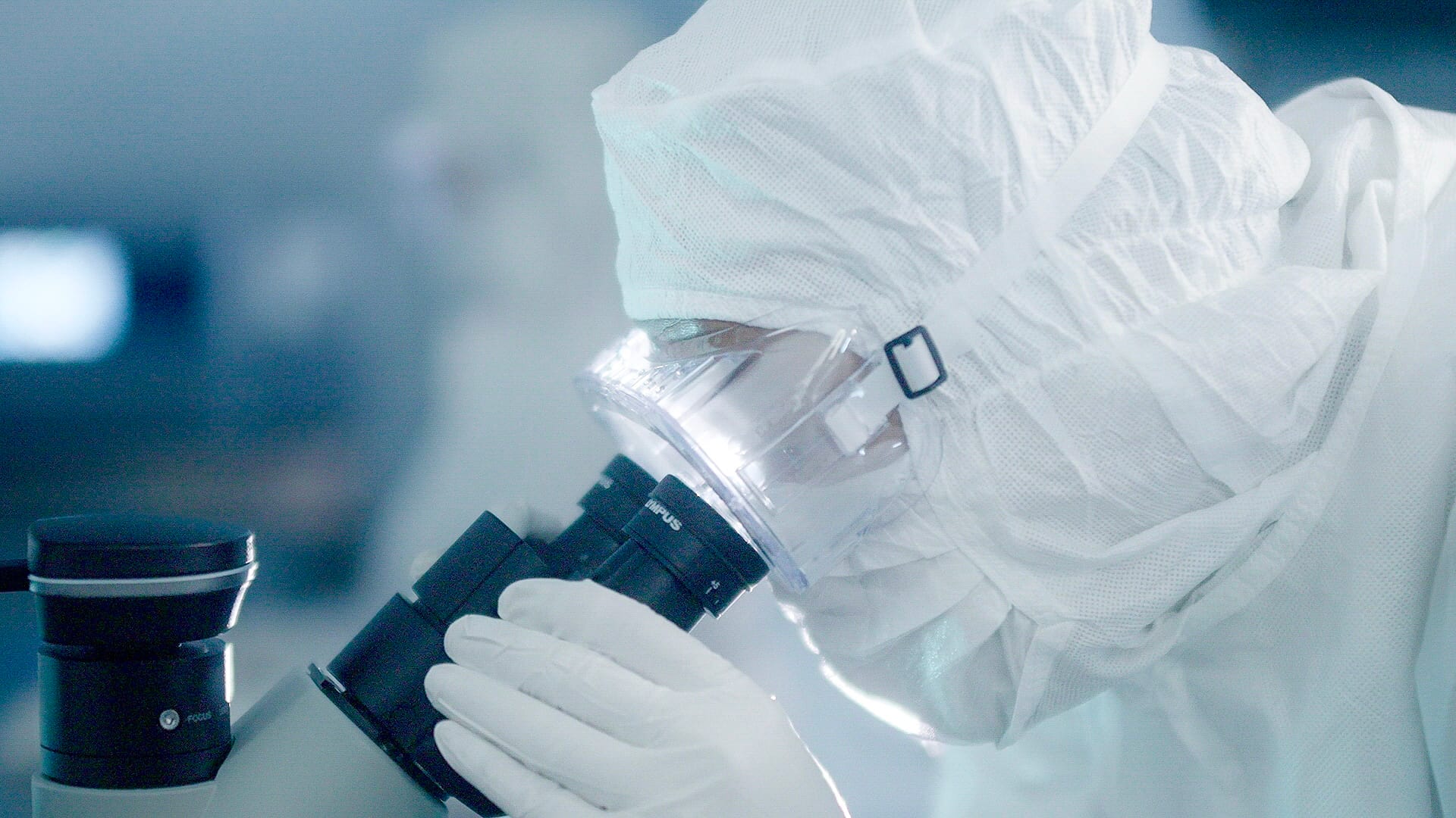 Fully gowned lab officer looking into a microscope.
