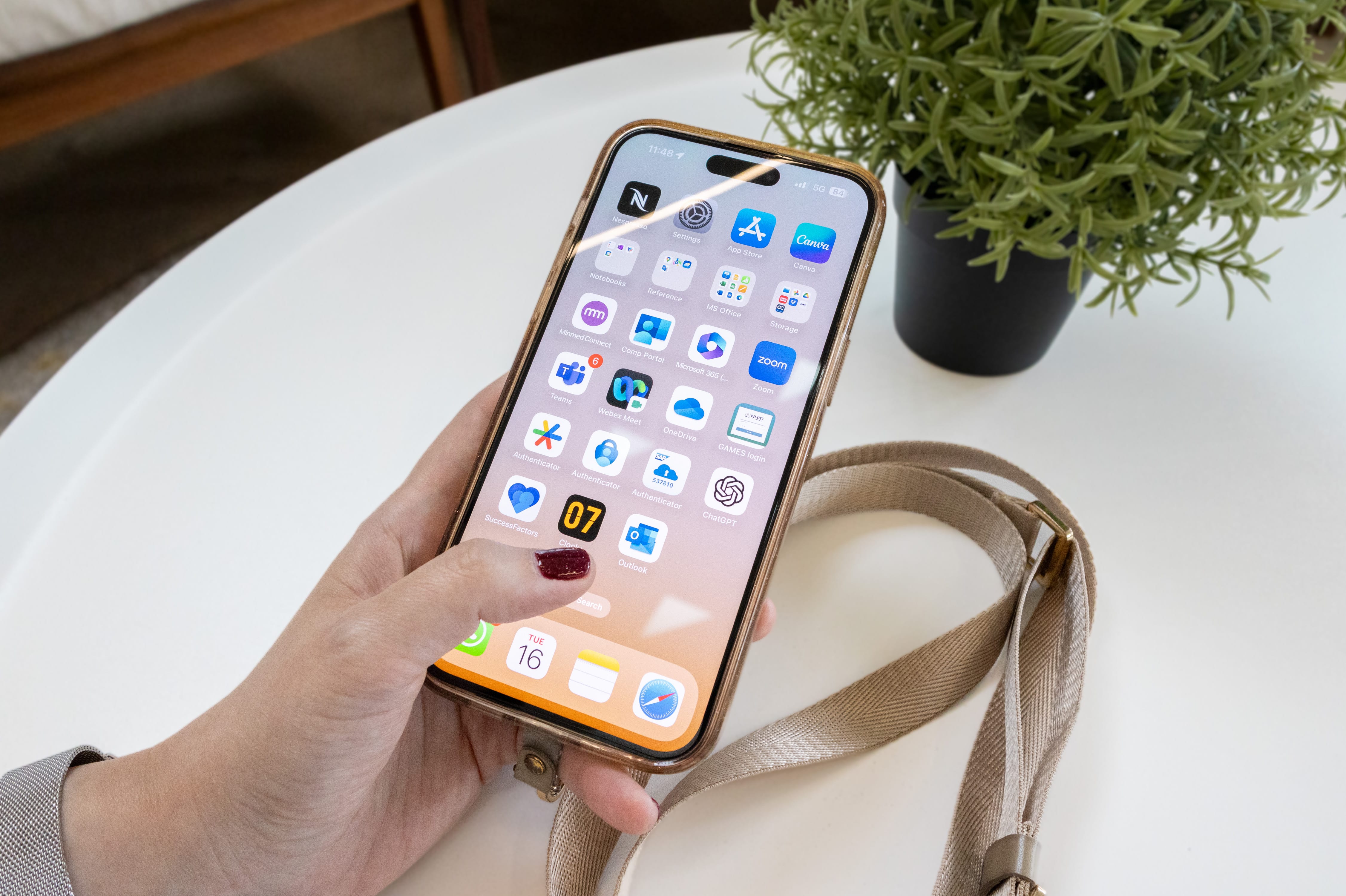 A hand with red nails holds an iPhone displaying a home screen with work apps on a white table near a plant.