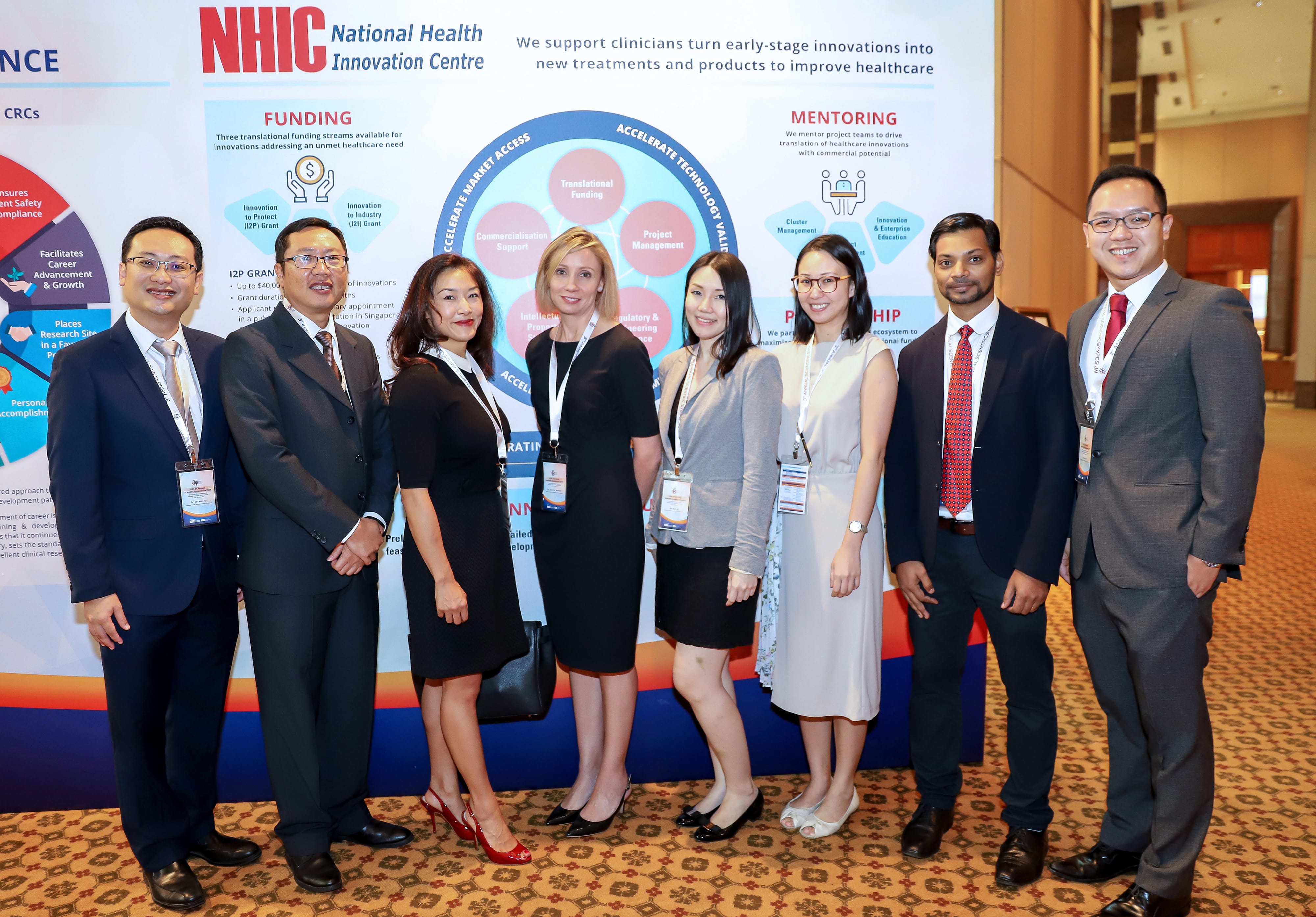 Seven professionals in formal attire pose smiling in front of an NHIC National Health Innovation Centre banner.
