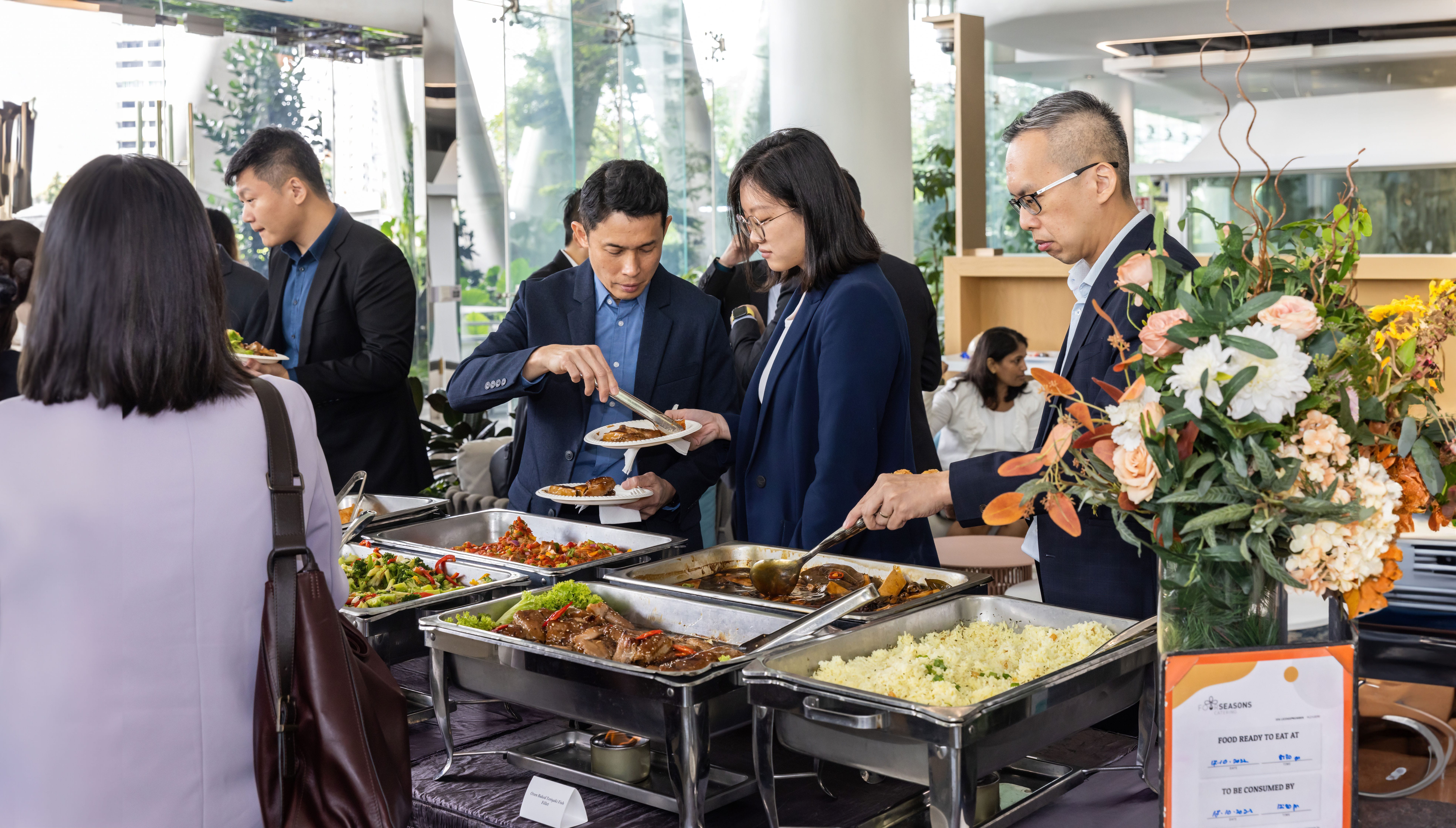 People in business attire serving themselves food from buffet chafing dishes with rice, meat, and vegetables.