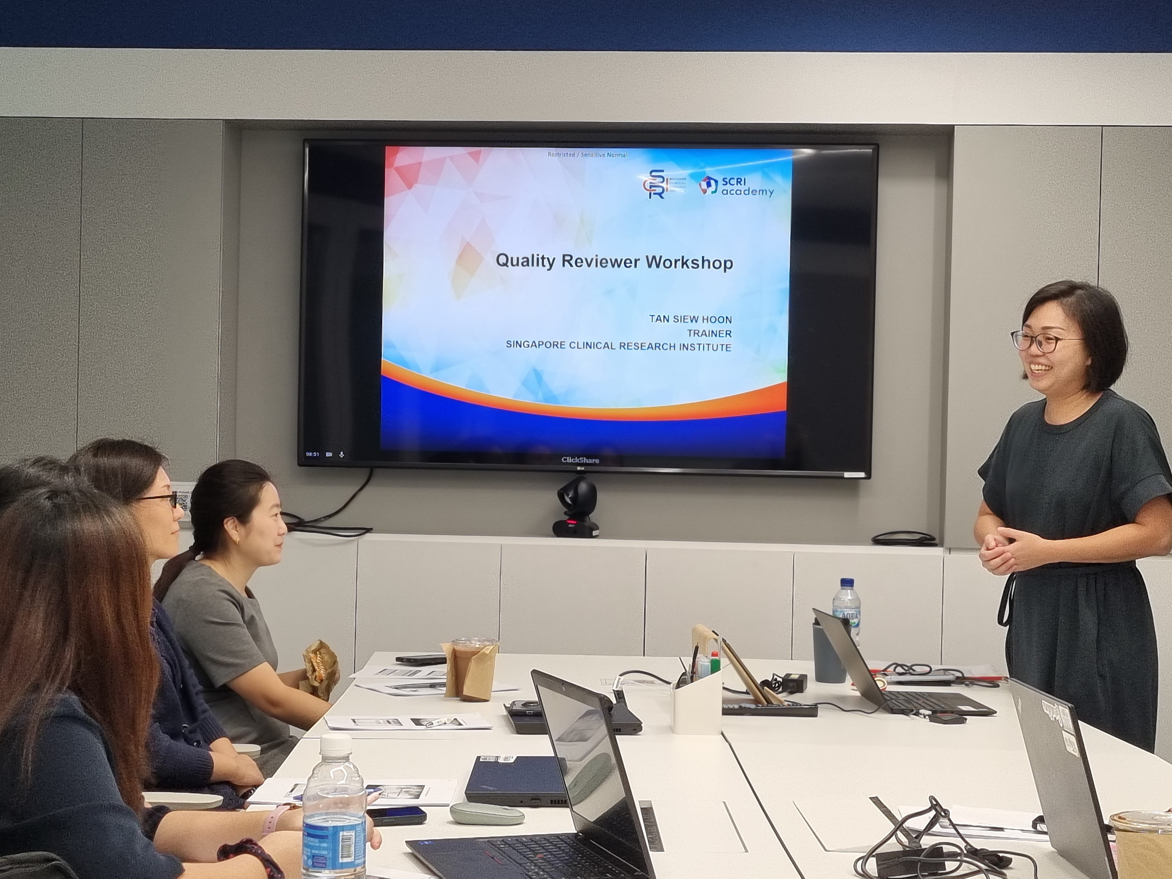 A trainer stands presenting a Quality Reviewer Workshop slide to two seated attendees in a conference room.