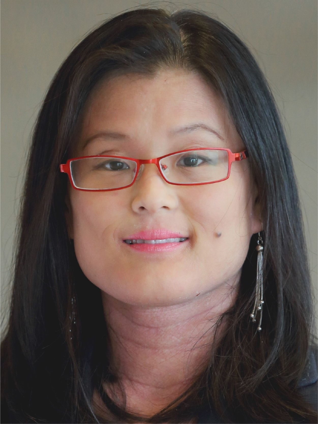 A woman with long dark hair, red-framed glasses, dangling earrings, and pink lips smiling at the camera.
