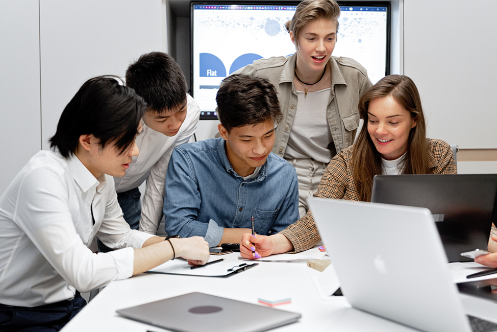 Five young people gathered around a table with laptops, collaborating on notes with a screen displaying slides behind them.