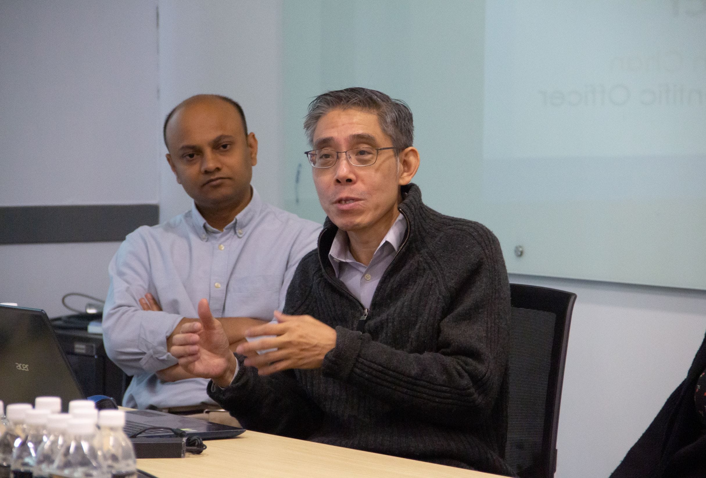 Two men sit at a conference table; one in a dark sweater and glasses speaks while the other in a blue shirt listens.