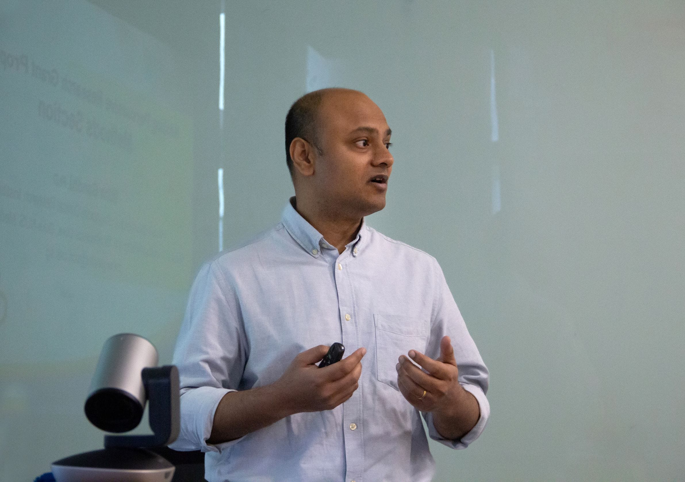 A man in a light blue shirt holds a clicker and gestures while presenting in front of a projected slide.