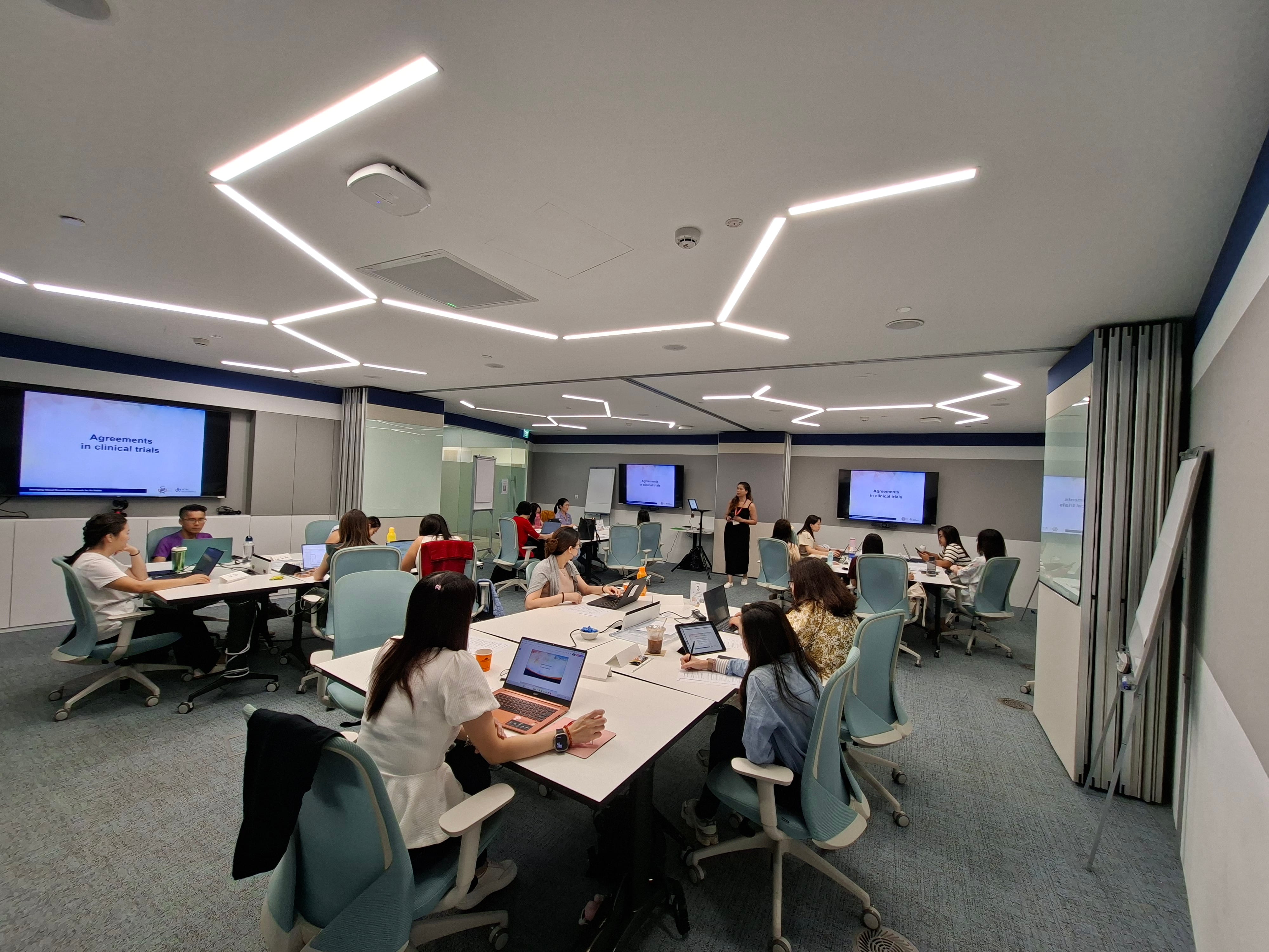 Instructor stands between two screens presenting slides to seated participants in a classroom with blue chairs.