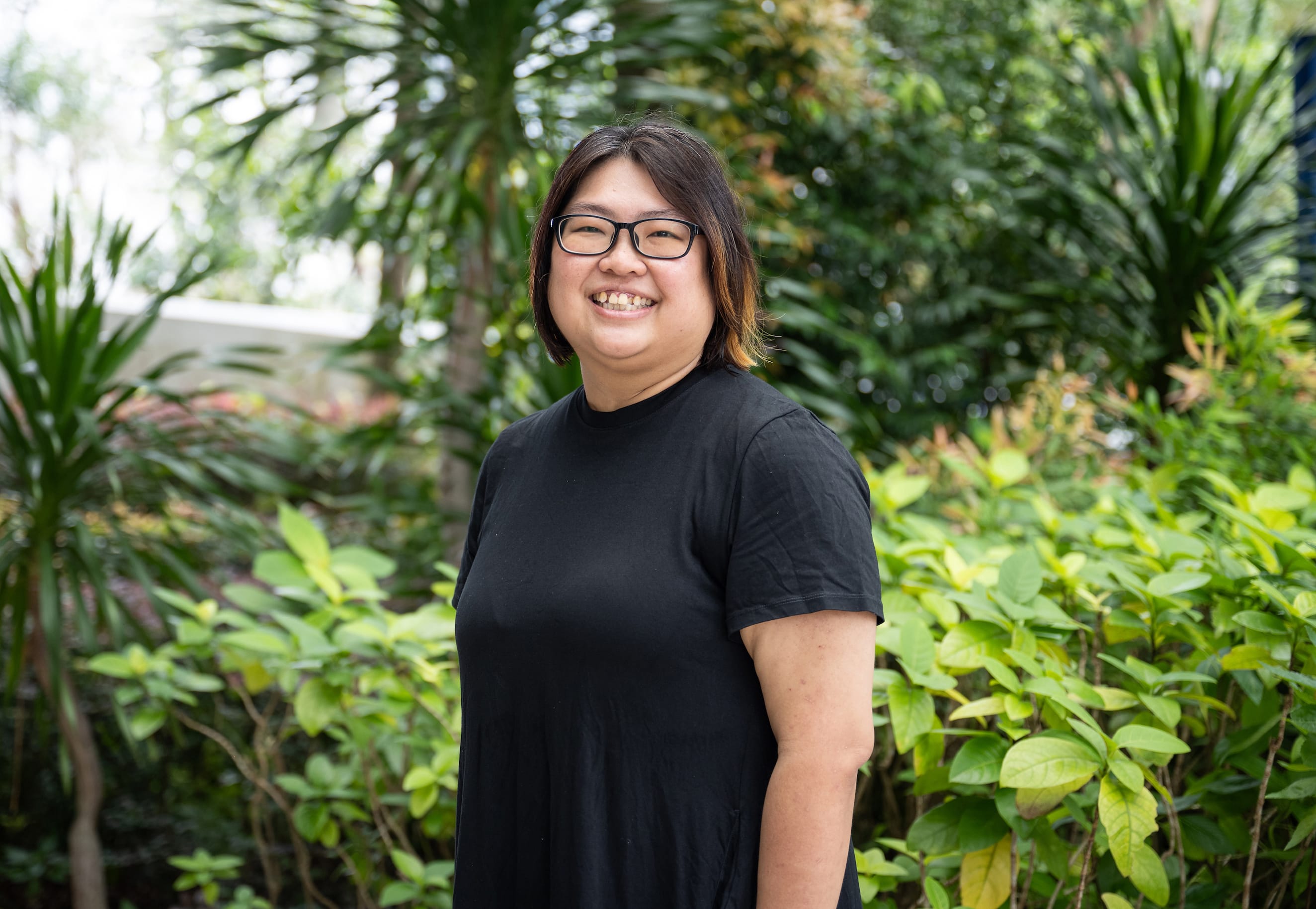 A smiling woman with glasses and short dark hair wearing a black t-shirt, standing outdoors among green tropical plants.