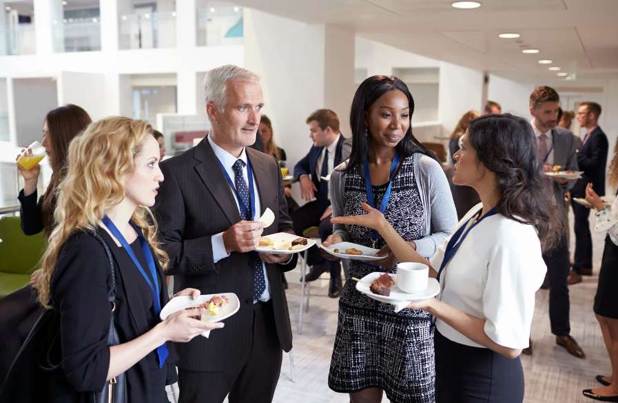 Four professionals wearing lanyards chat while holding plates of food at an indoor networking event.