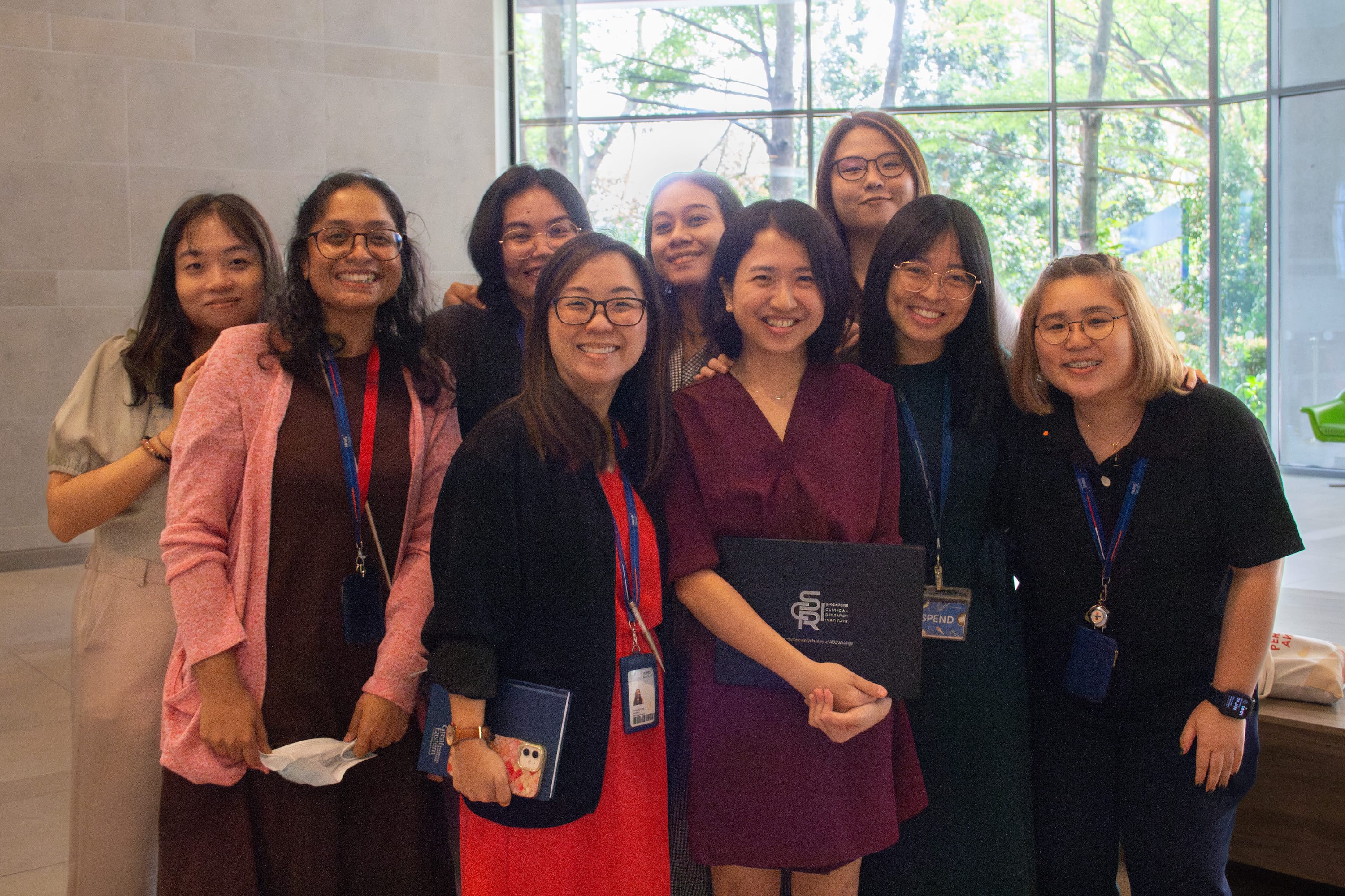 A group of nine smiling women wearing lanyards pose together indoors in front of a large window with trees outside.