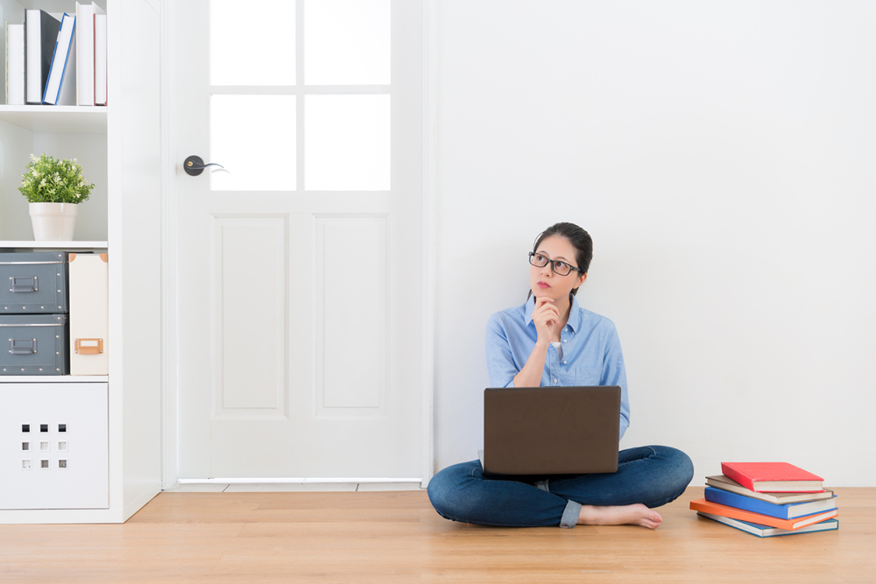 Woman with glasses sits cross-legged on a wood floor with a laptop, looking up thoughtfully, books stacked nearby.