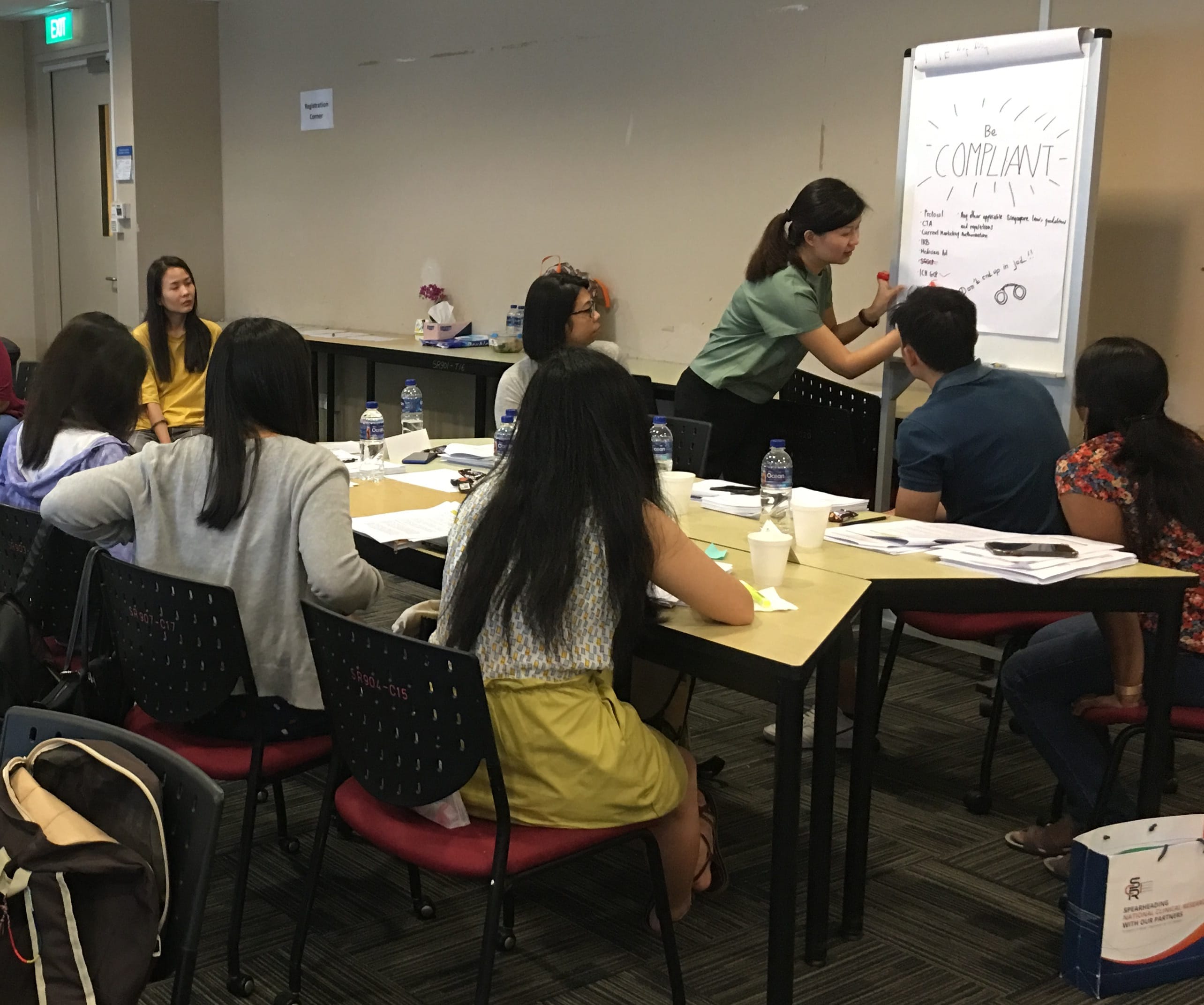 A woman writes "Be Compliant" on a flipchart while several people sit around tables in a classroom workshop.