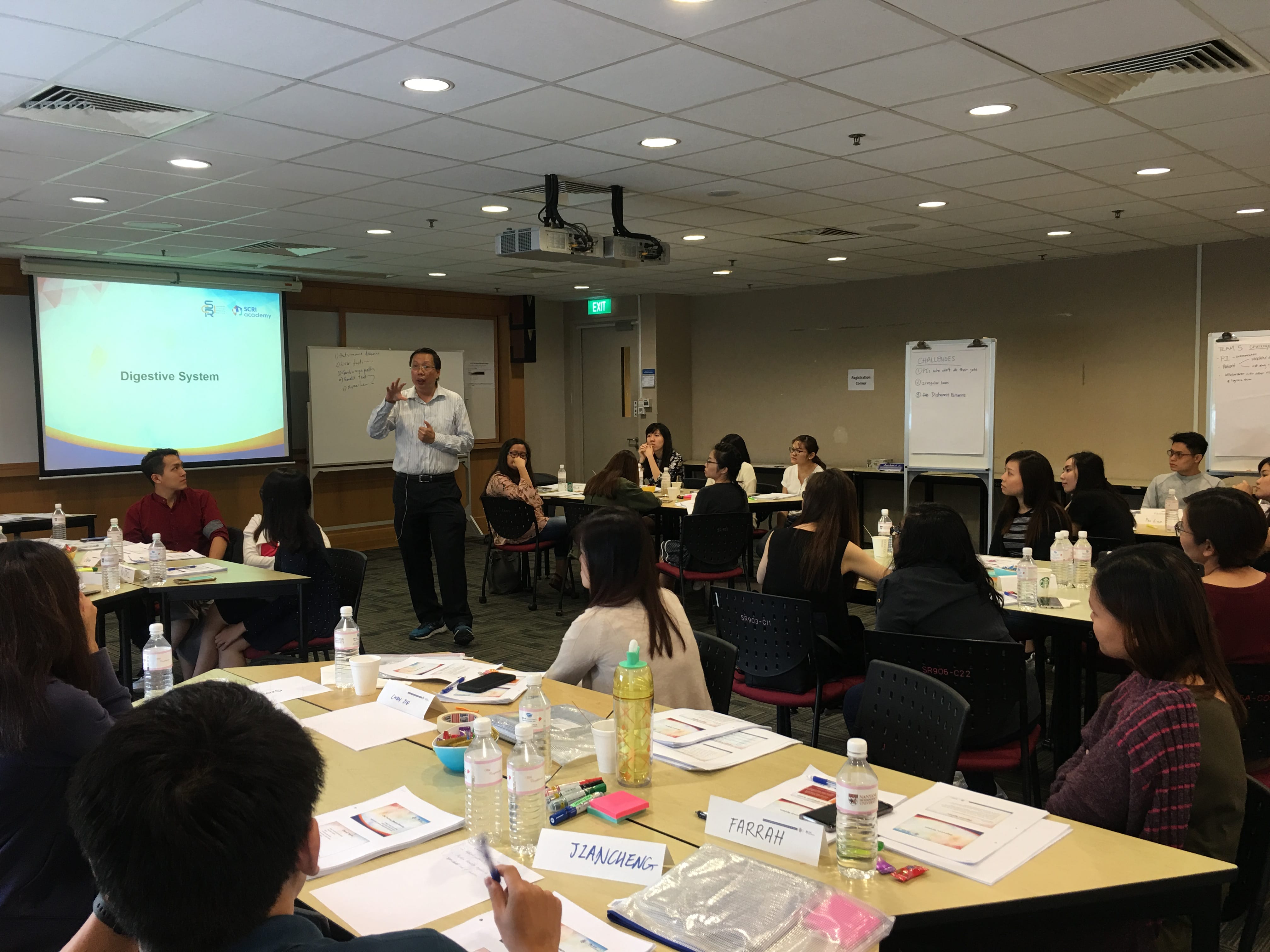 A man stands presenting a Digestive System slide to students seated at tables with name cards in a classroom.