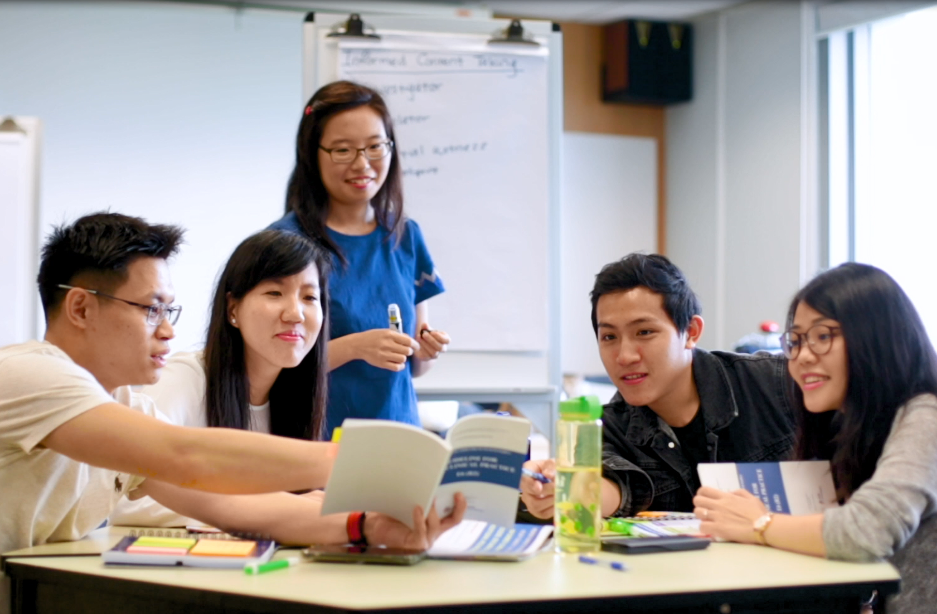 Five students gathered around a table with books, one standing near a whiteboard during a group study session.