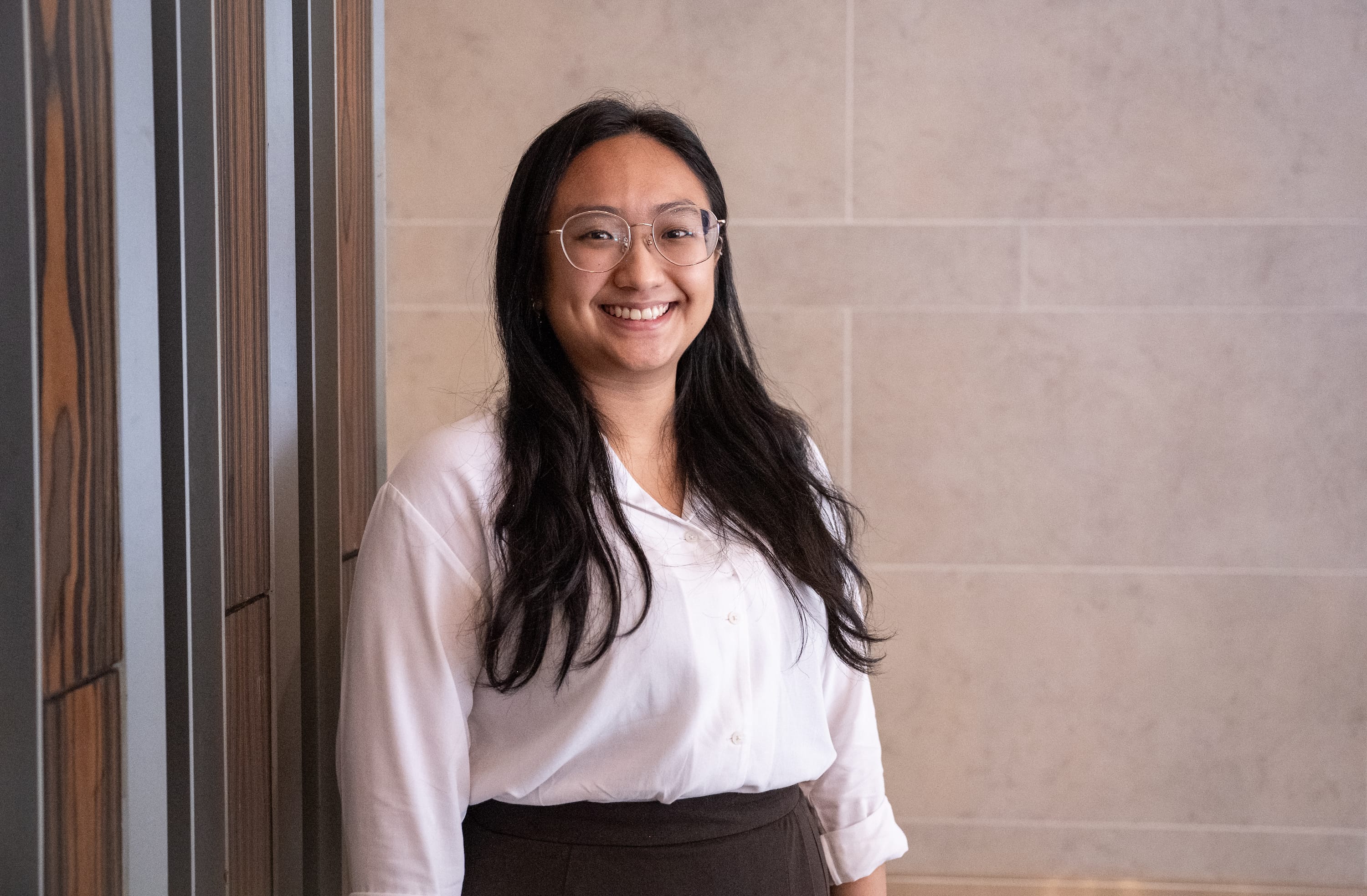 Smiling person with long dark hair and glasses, wearing a white blouse, standing by a tiled wall.