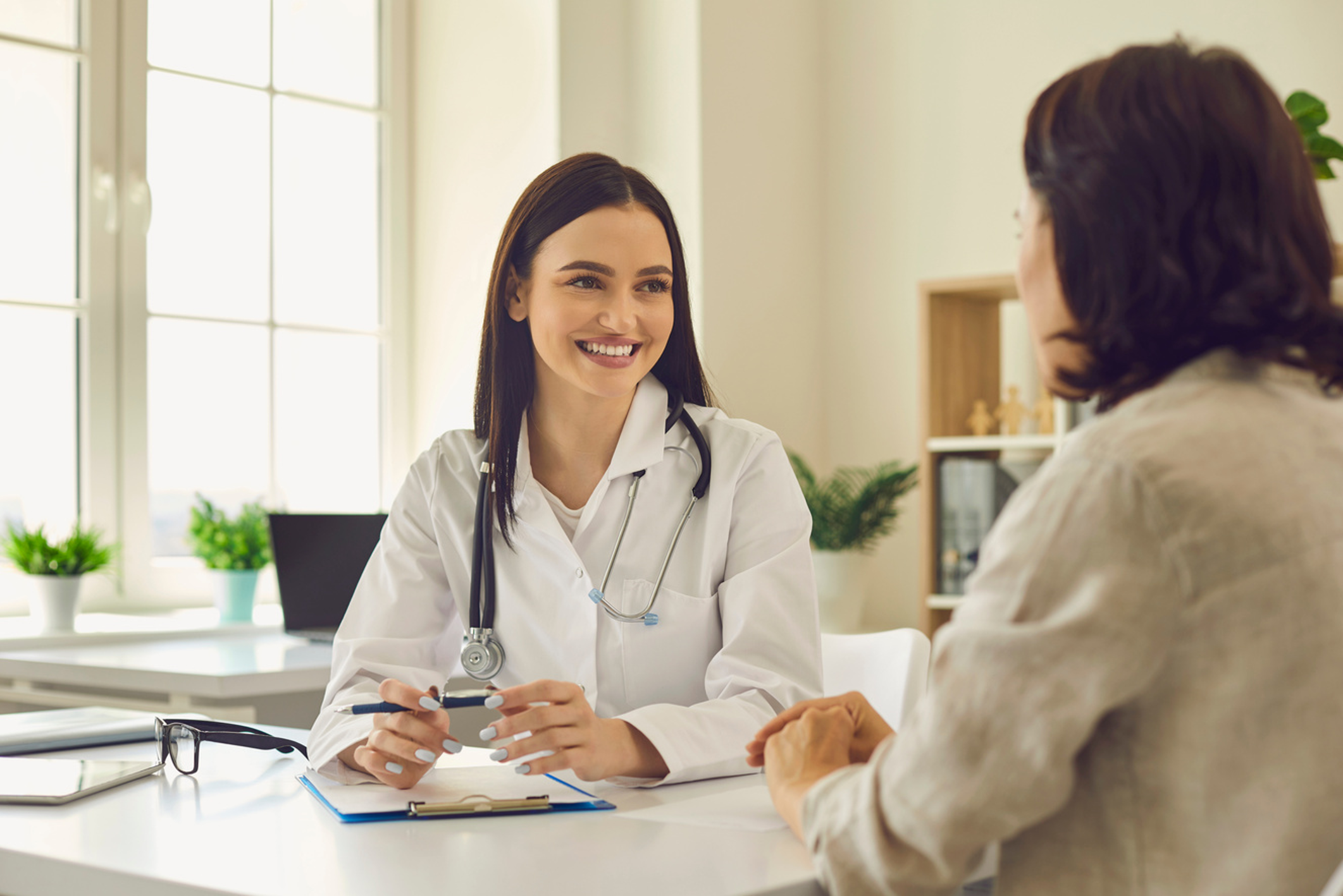 A smiling female doctor in a white coat with stethoscope consults a patient at a desk in a bright office.