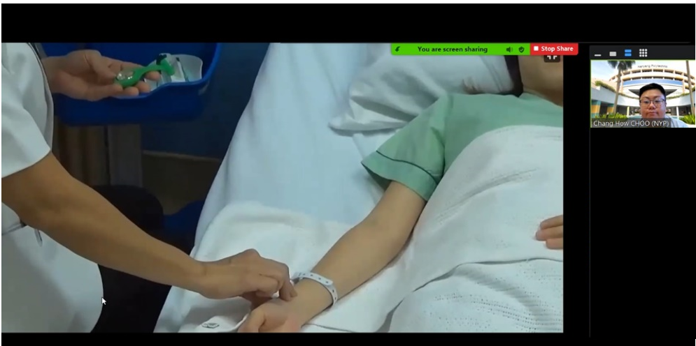 A video call screen showing a nurse attending to a patient lying in a hospital bed with a wrist ID band.
