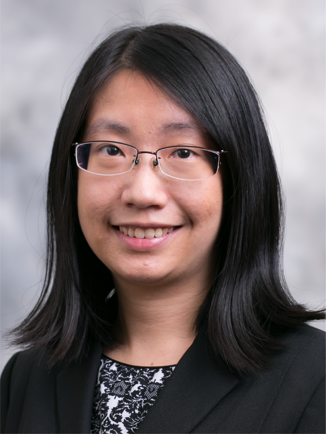 A smiling woman with long black hair and thin-framed glasses wearing a black blazer and patterned top.
