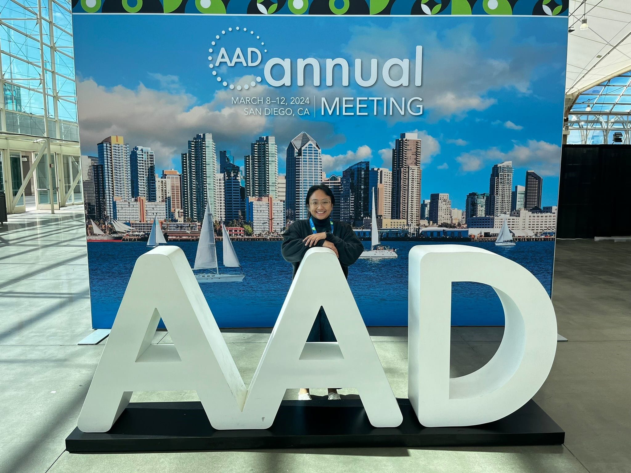 Person smiling behind large AAD letters at the AAD Annual Meeting 2024 banner in San Diego, CA.