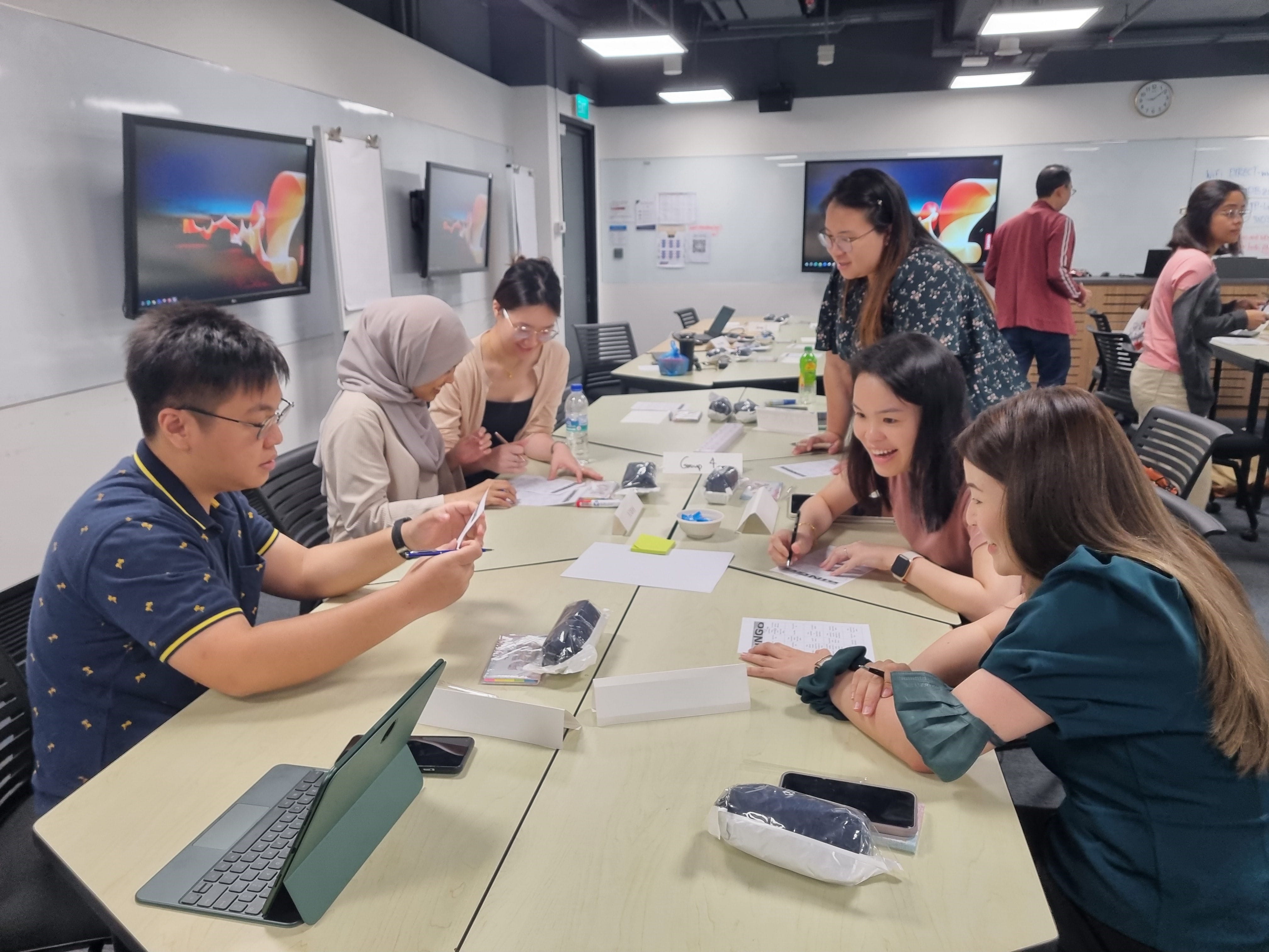 Six people sit around a table in a classroom, writing on papers, with screens and whiteboards on the walls.