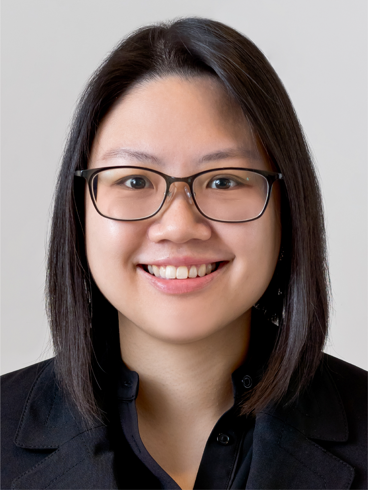 A young woman with straight dark hair, rectangular glasses, and a black top smiles against a gray background.