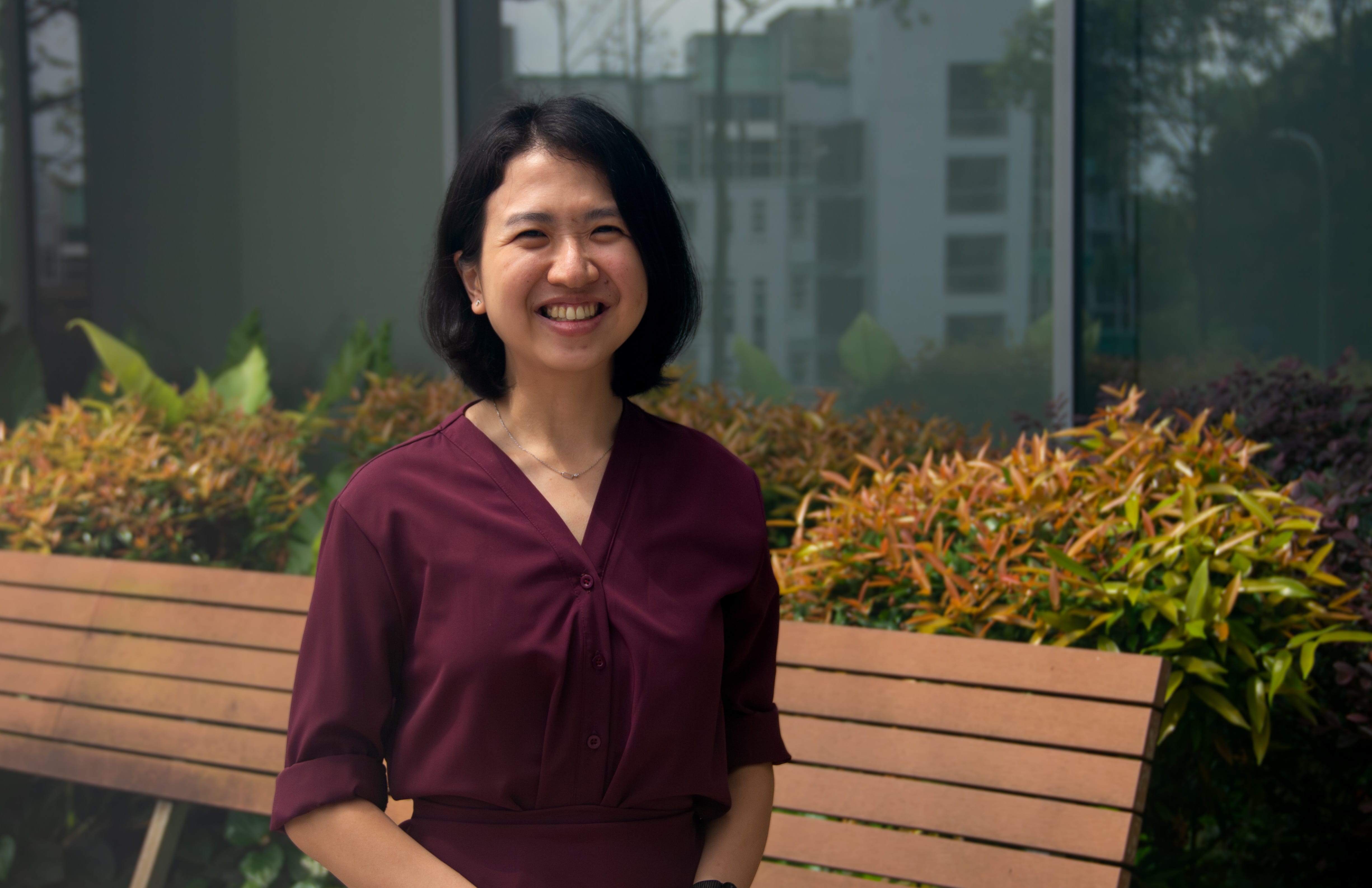 A smiling woman with short black hair in a dark red blouse stands outdoors near a wooden bench and colorful shrubs.