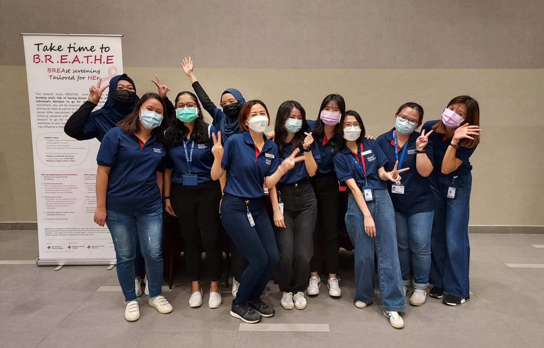Group of masked women in blue NUHS uniforms posing near a B.R.E.A.T.H.E breast screening awareness banner.