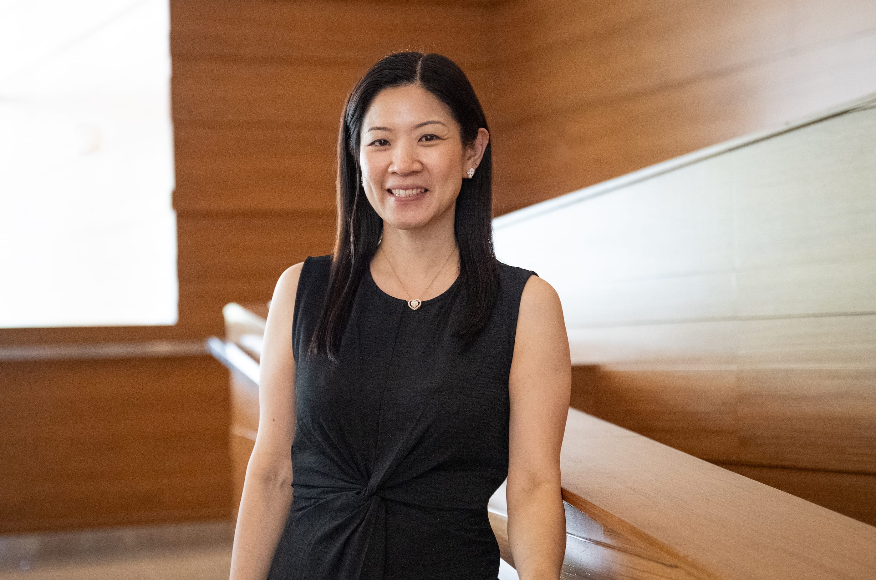 Smiling woman in a sleeveless black dress leans on a wooden railing in a bright, modern interior.