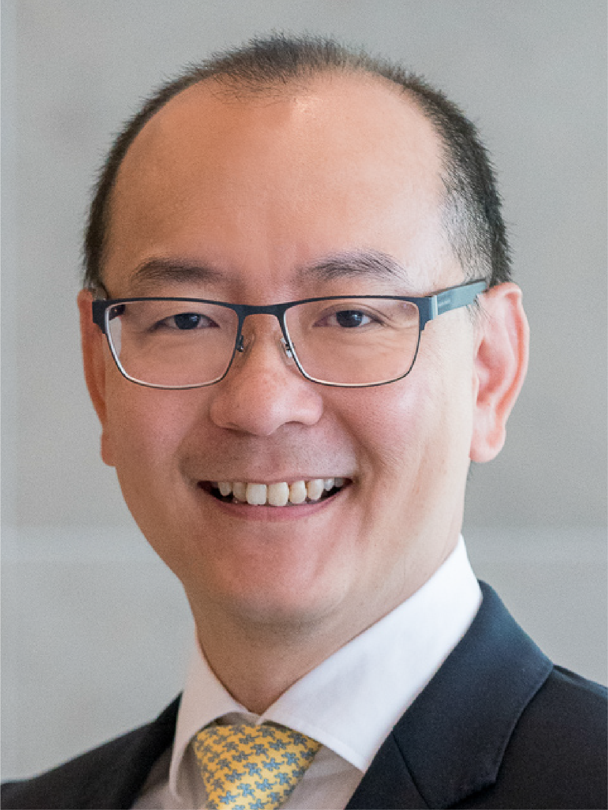 A smiling middle-aged man wearing dark-framed glasses, a dark suit, white shirt, and yellow patterned tie.