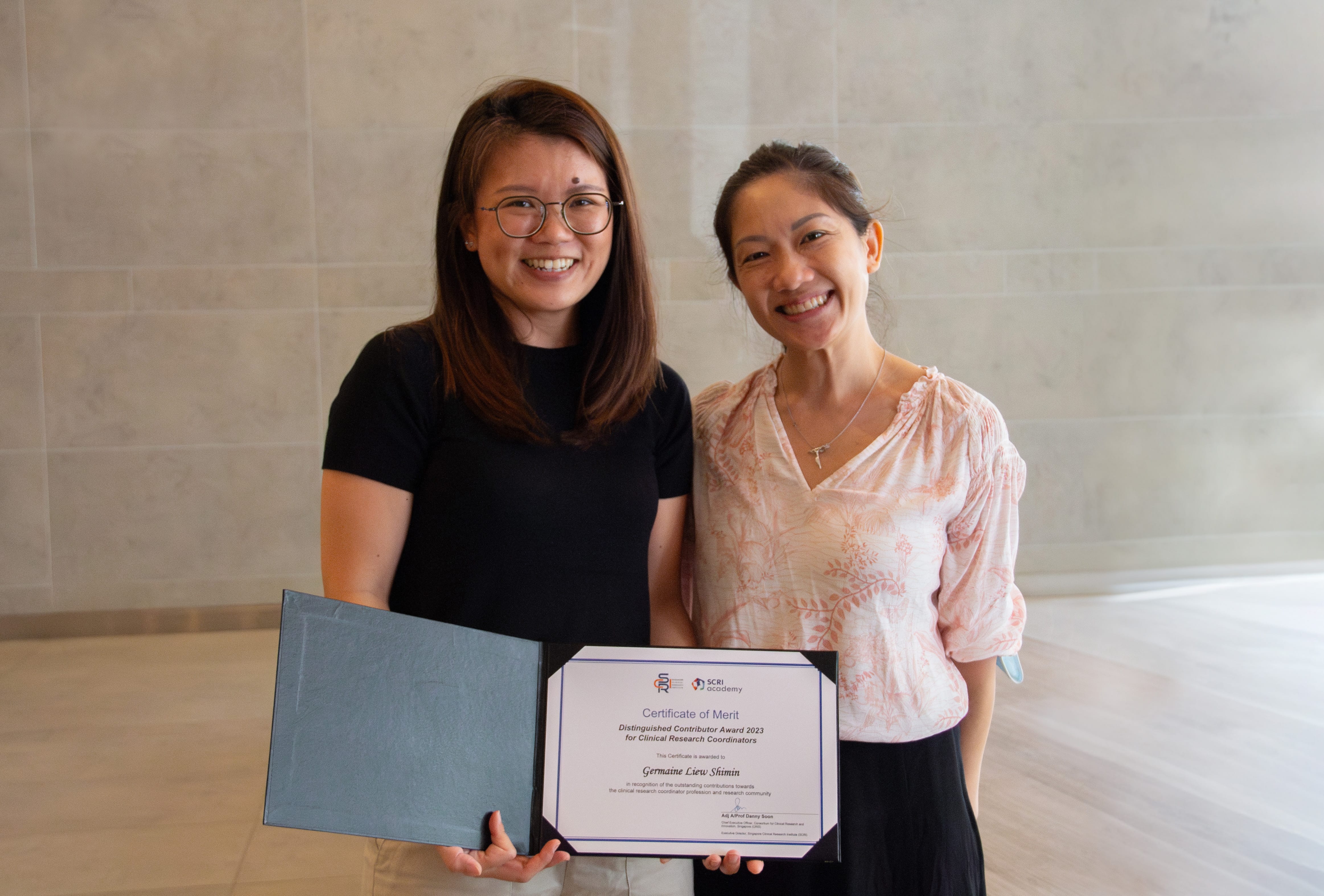 Two women smile, one holding an open folder displaying a certificate, standing against a light tiled wall.