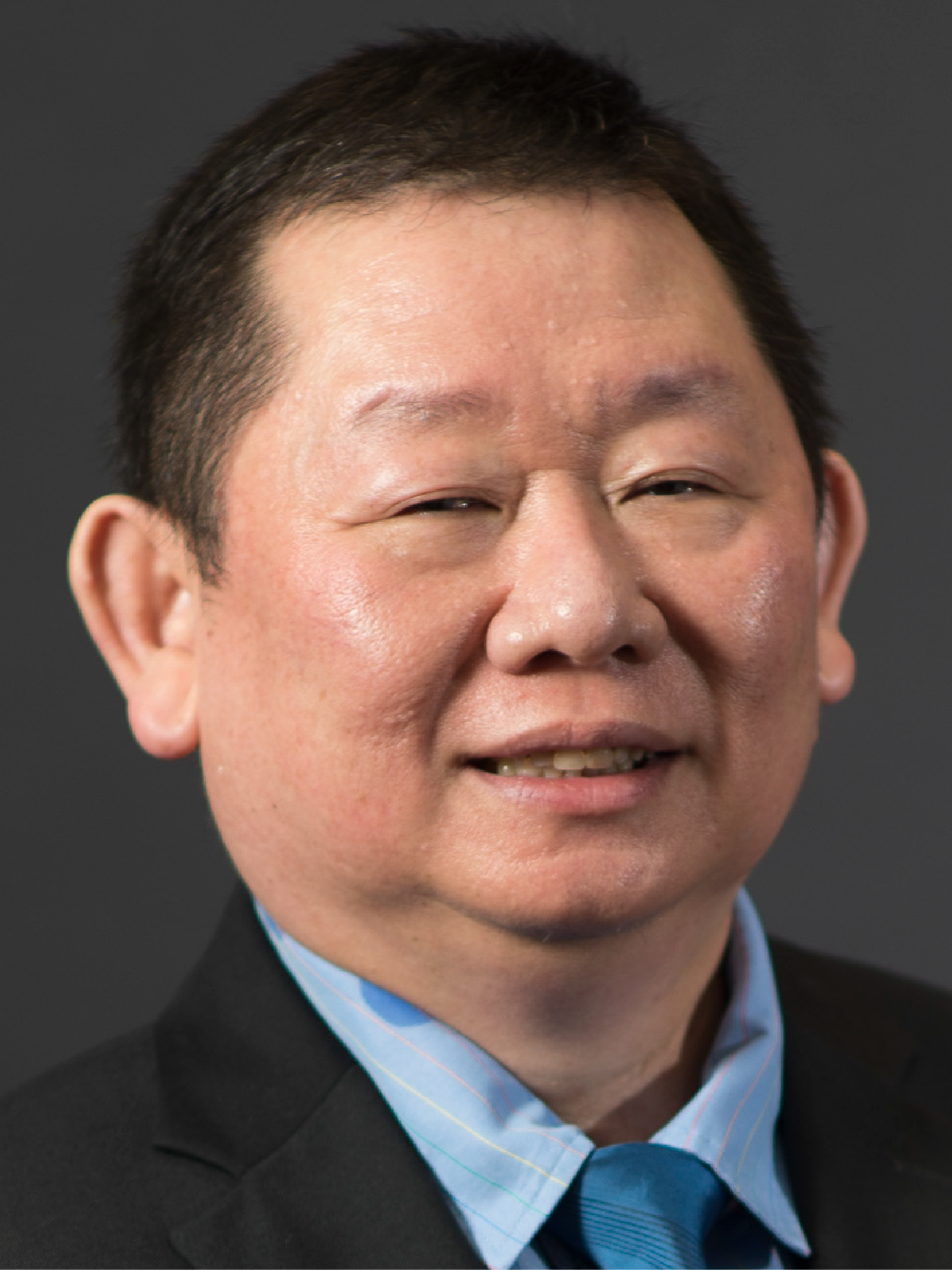 A middle-aged man smiling, wearing a dark suit, light blue striped shirt, and teal tie, against a gray background.