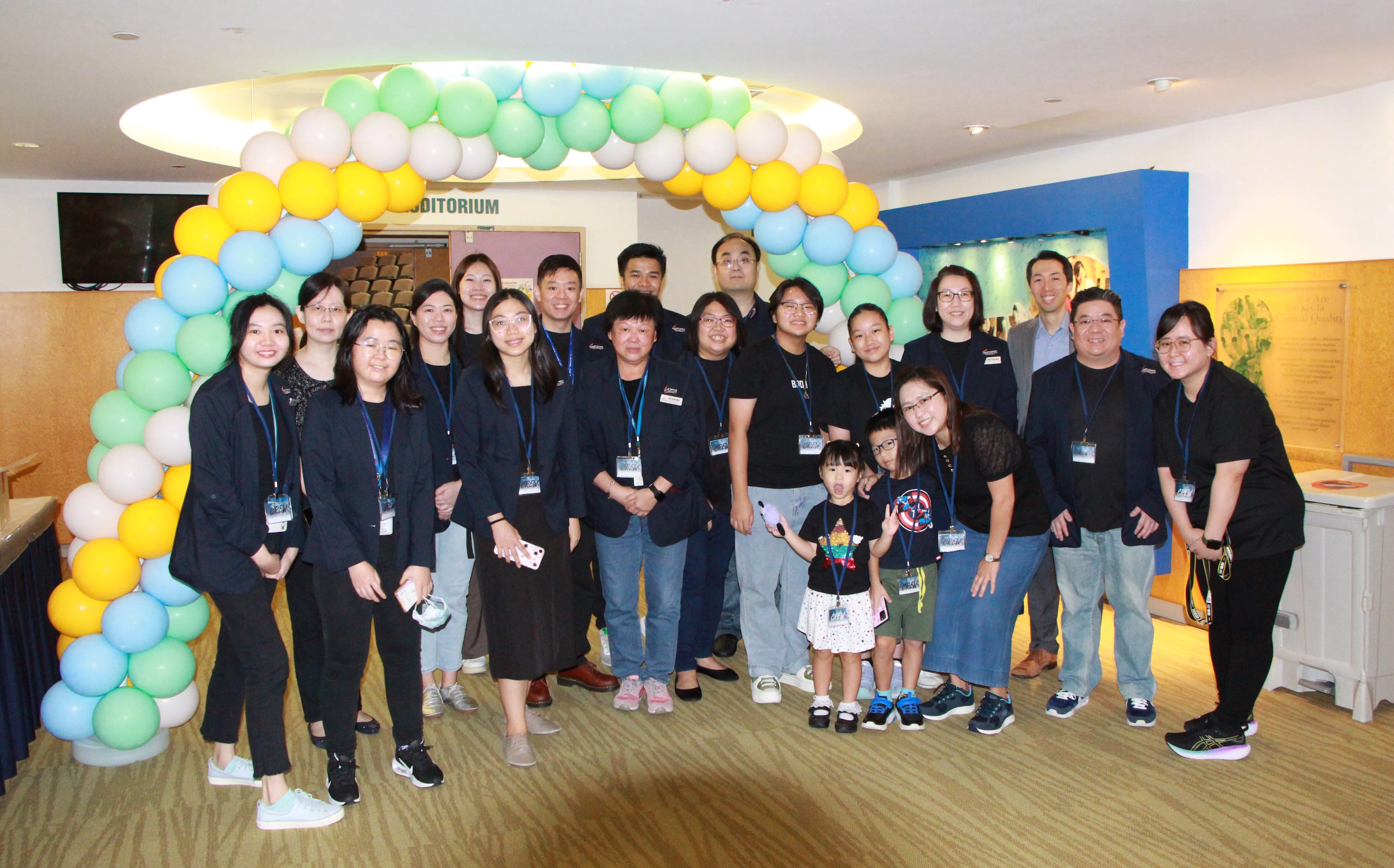 Group of adults and two children posing under a pastel balloon arch indoors, most wearing black and lanyards.