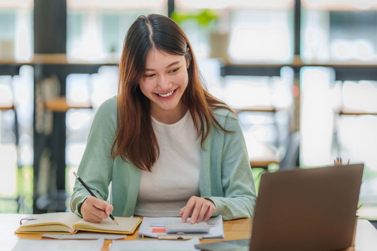 Young woman smiling while writing in a notebook at a desk with an open laptop nearby.