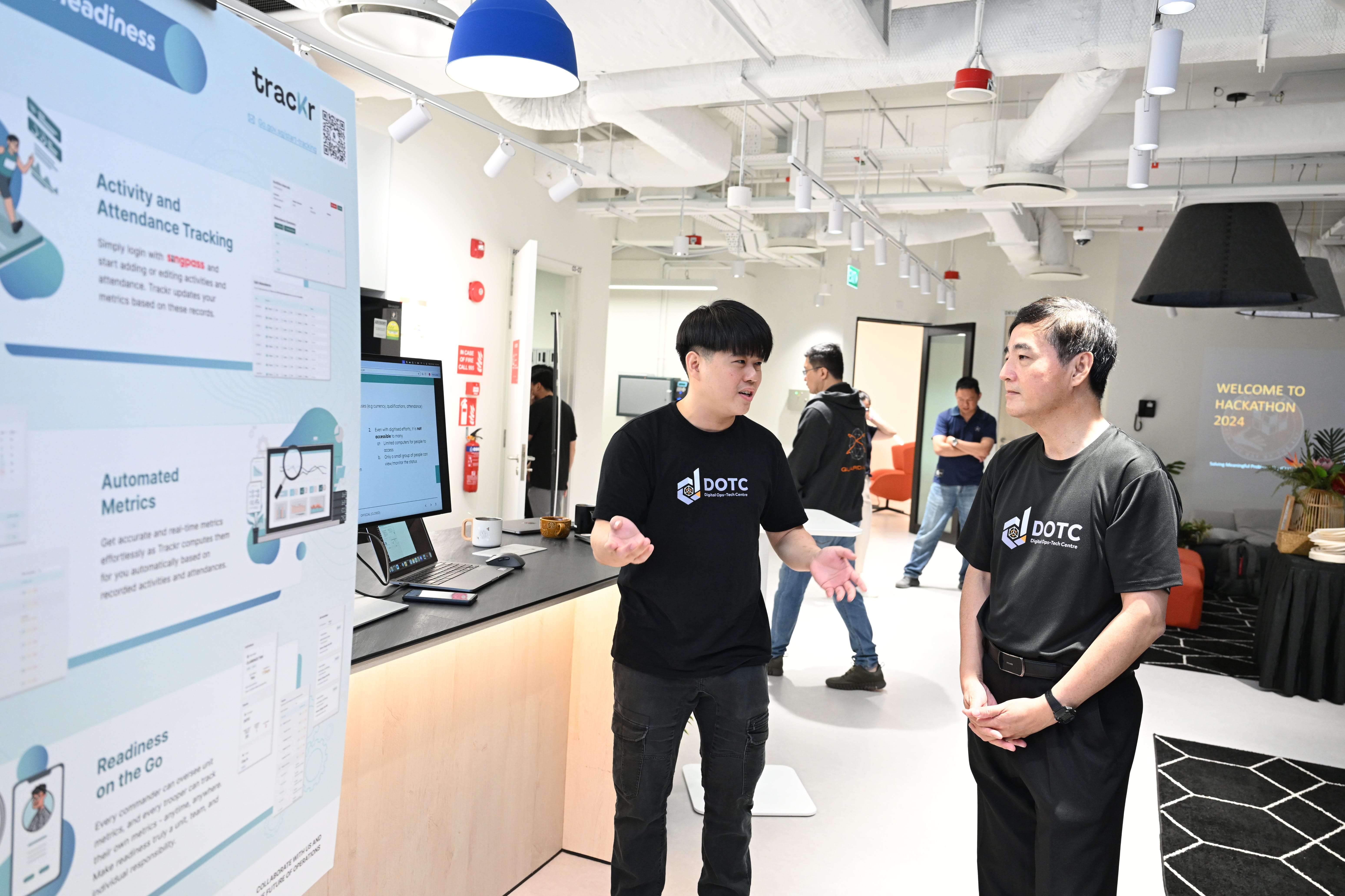 Two men in DOTC shirts discuss a TracKr activity tracking poster at Hackathon 2024 in a modern office.