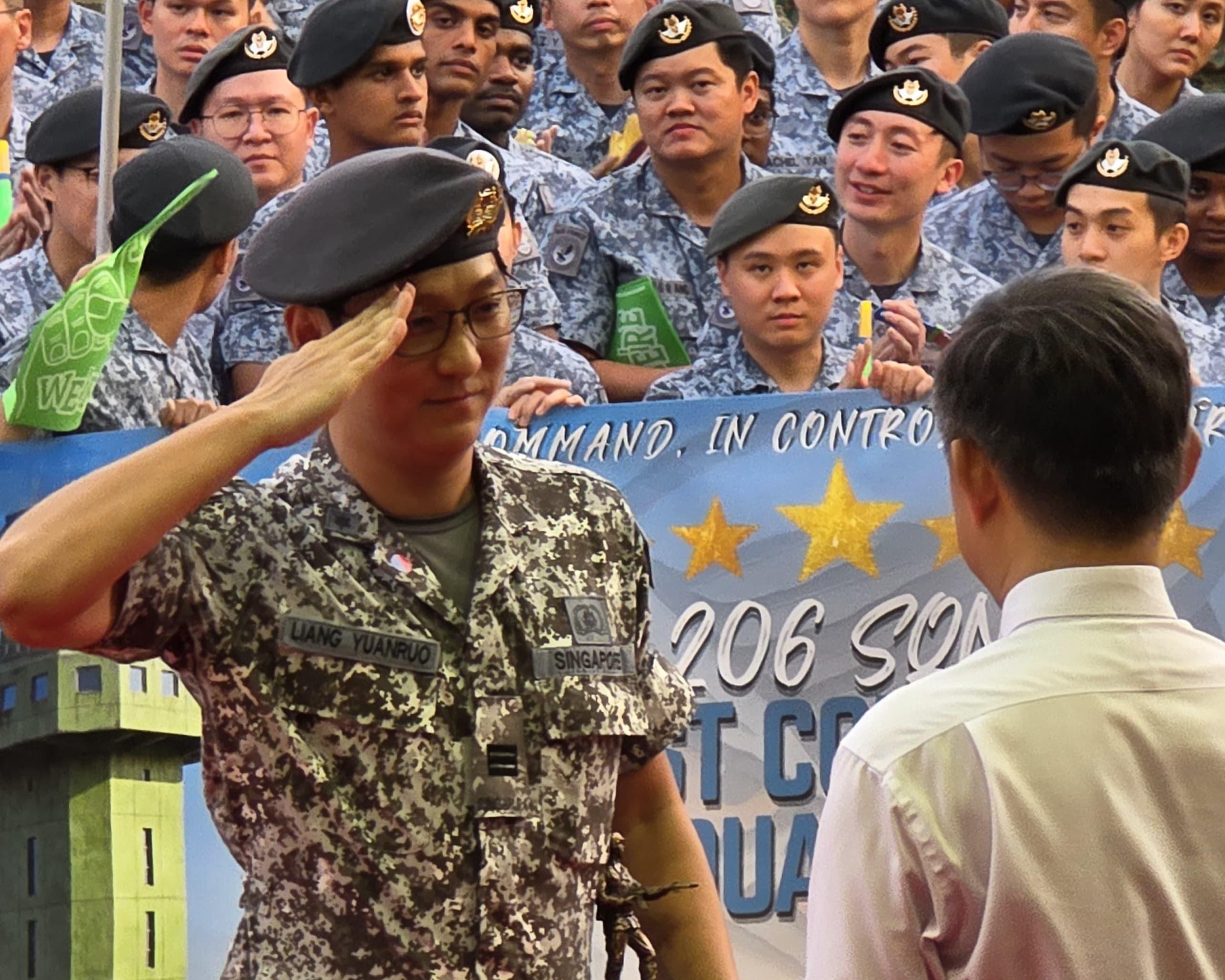 A medium shot of an East Asian man in a pixelated Singapore military camouflage uniform and black beret, holding a small trophy while rendering a formal salute to an official in a white shirt, with a crowd of soldiers in uniform standing in the background.