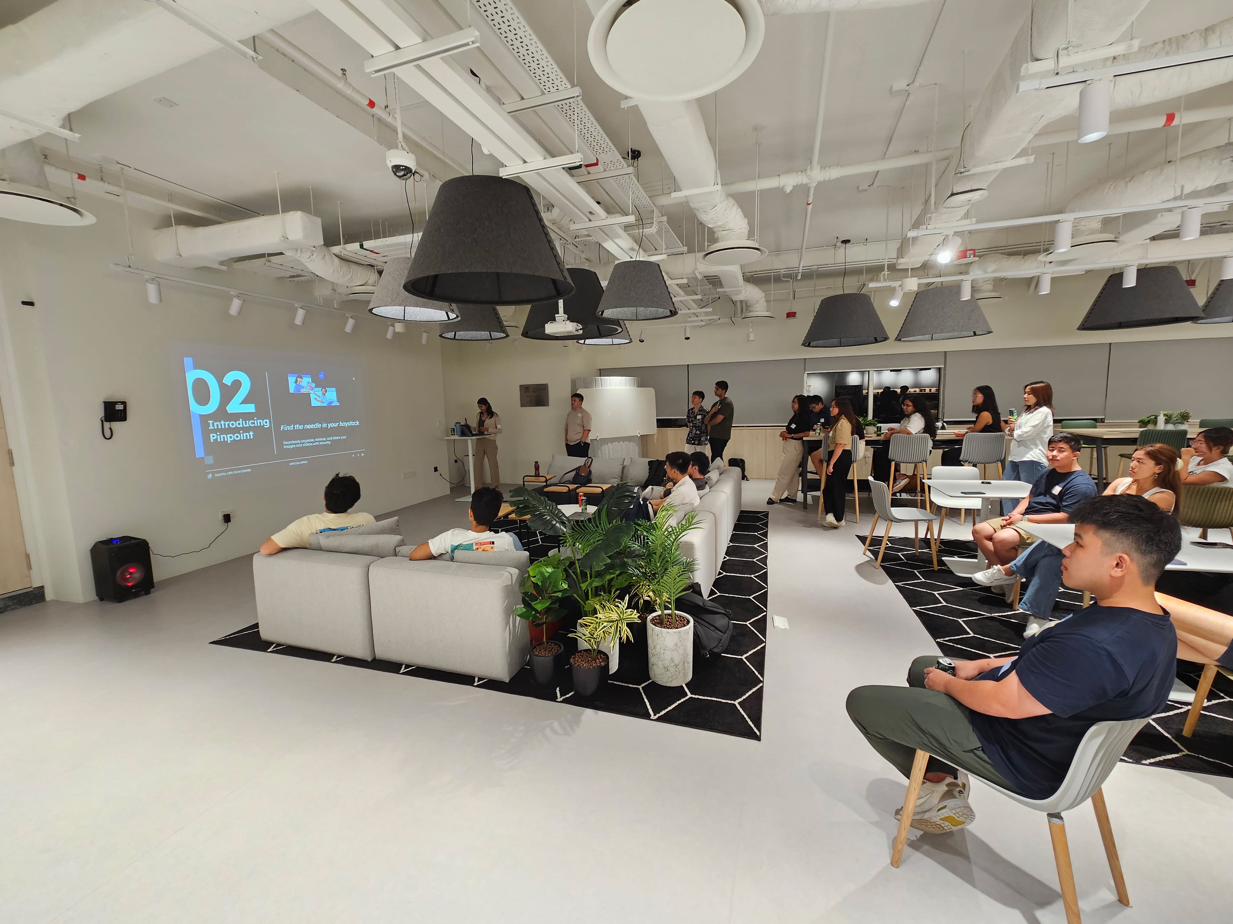 People sit on sofas and chairs in a modern lounge watching a presentation projected on a wall.