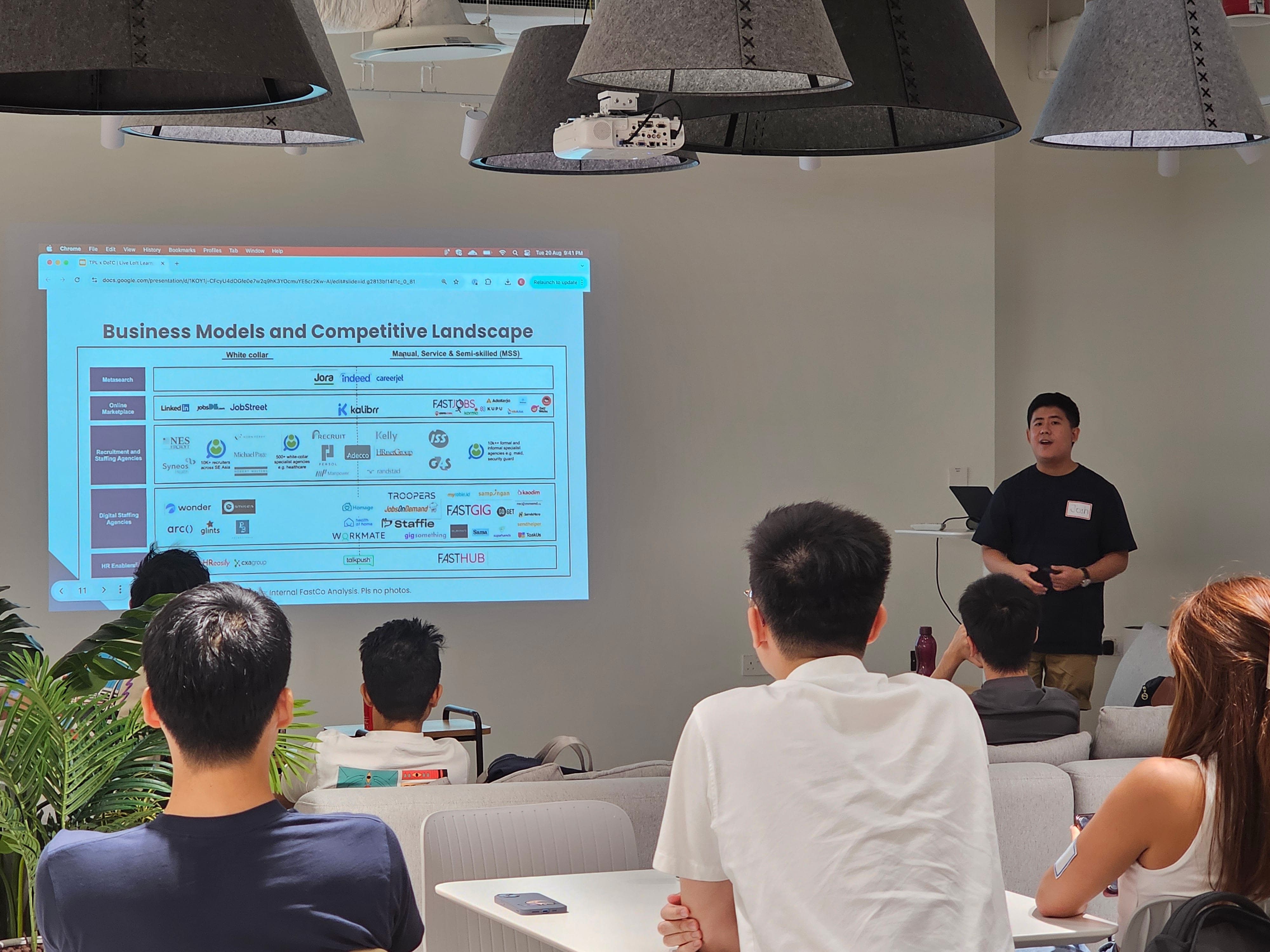 A man presents a slide titled "Business Models and Competitive Landscape" to a seated audience in a modern room.